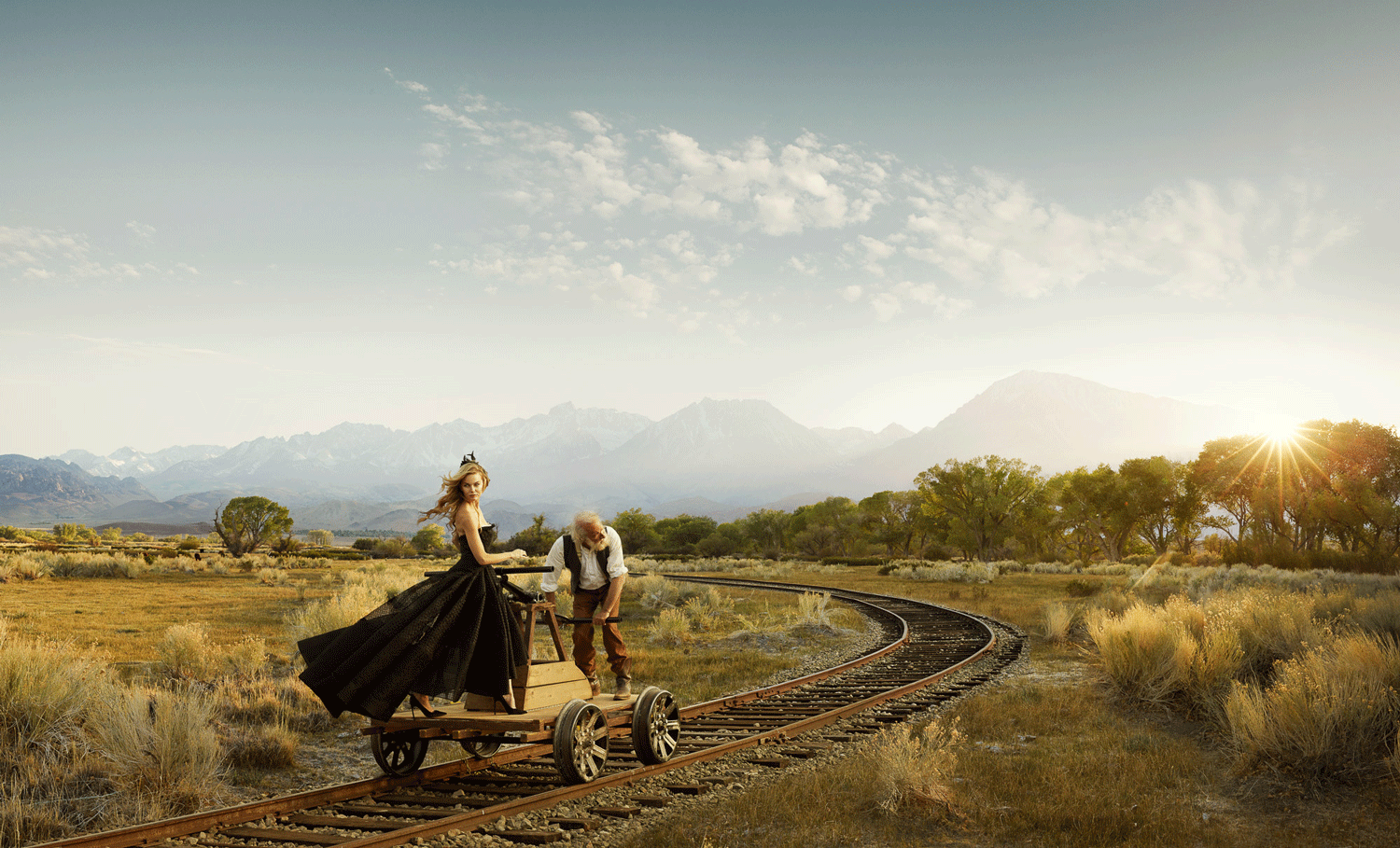 A surreal scene of a young woman in a black Victorian dress and an elderly man on a wooden train cart on railroad tracks in a desert landscape with mountains in the background during sunset.