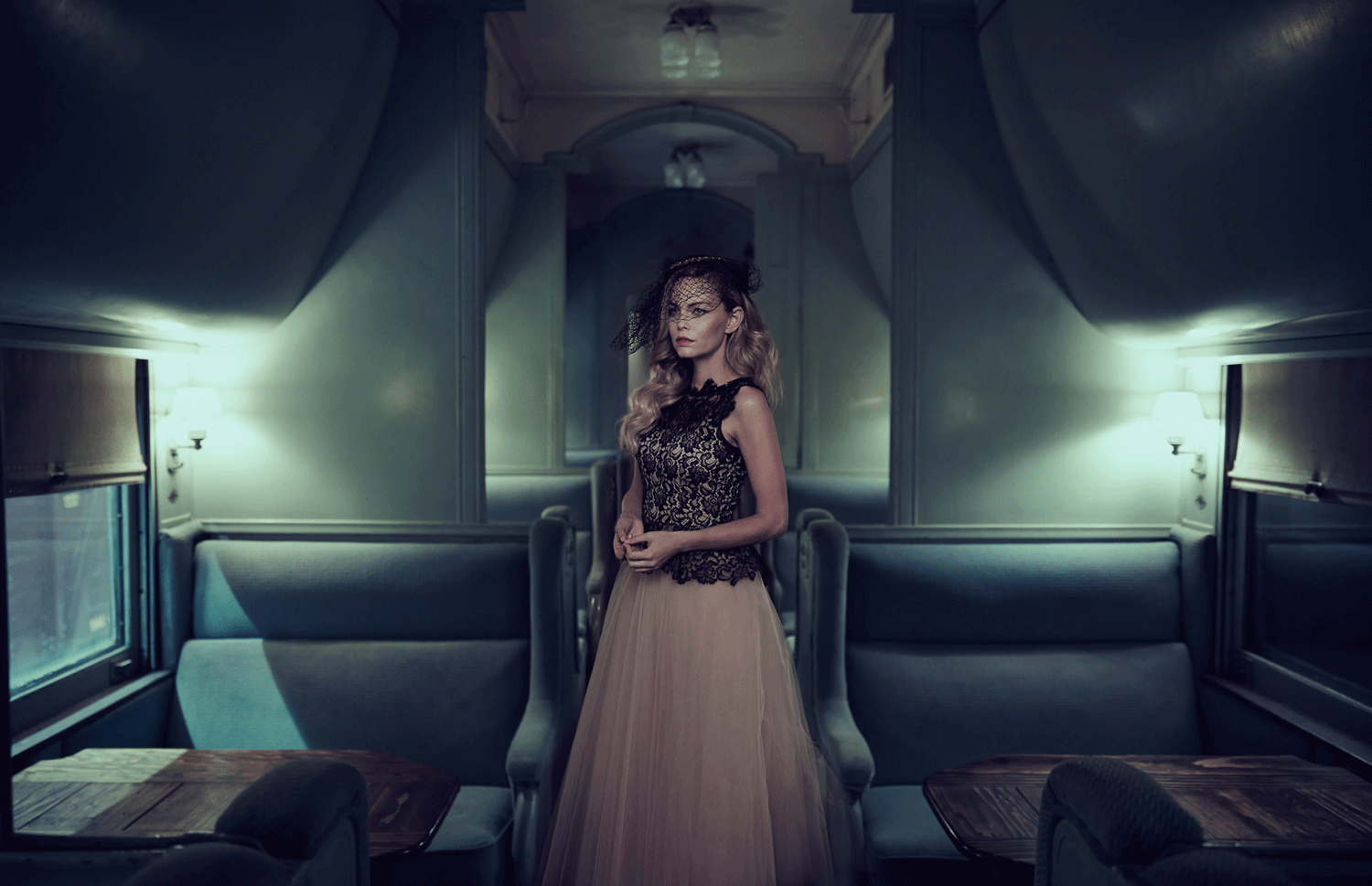 A woman in an elegant black lace top and beige tulle skirt stands inside a vintage train carriage, wearing a black hat with a veil, illuminated by soft interior lighting.