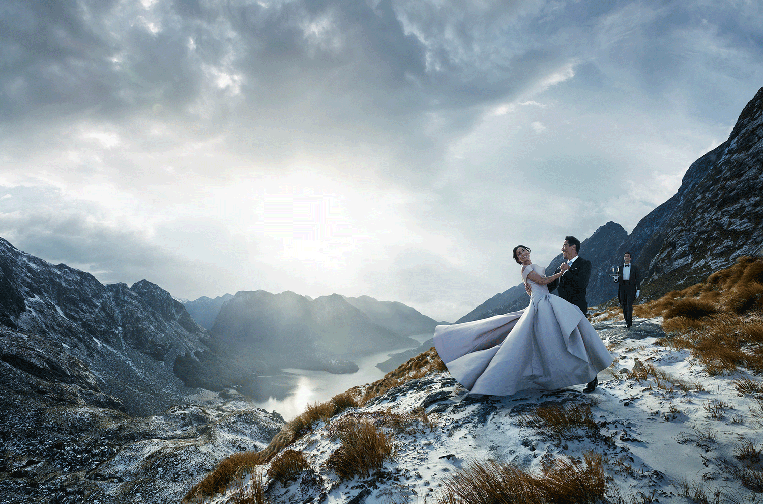 A bride and groom dancing on a snowy mountain landscape with a lake and mountain range in the background, with a waiter in a tuxedo carrying drinks in the distance.