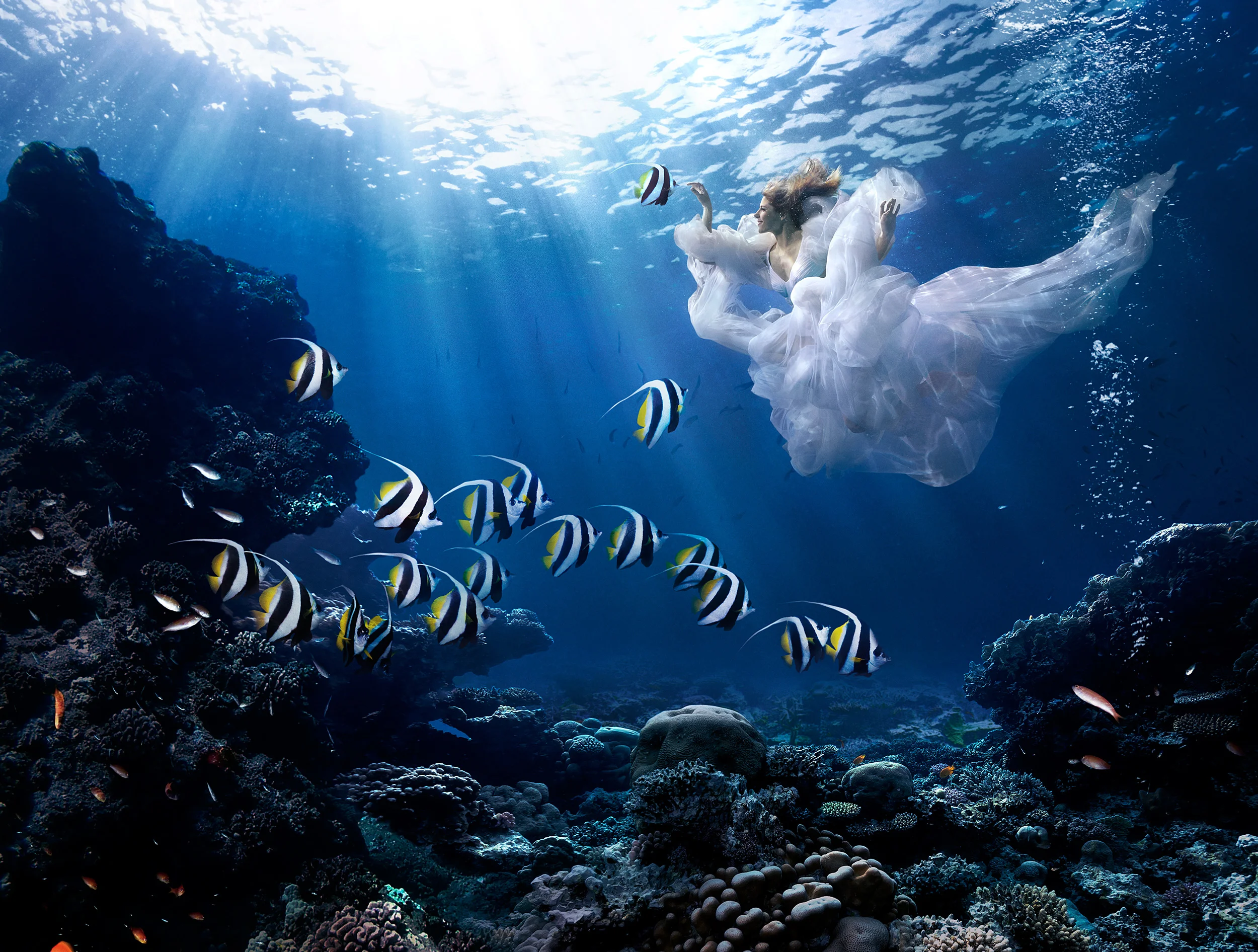 A woman in a flowing white dress swimming underwater near coral reefs and colorful fish, with light rays shining through the water.