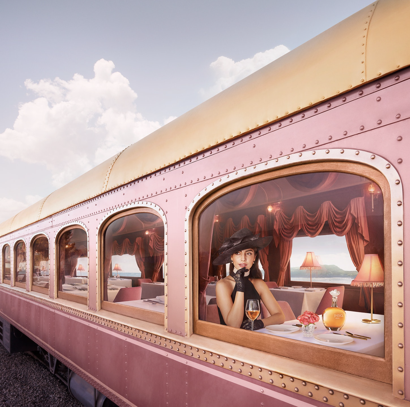 A woman dressed in vintage style sitting in a pink dining car with pink drapes, a lamp, a flower vase, and a glass of rosé wine, looking out the window at a scenic view.