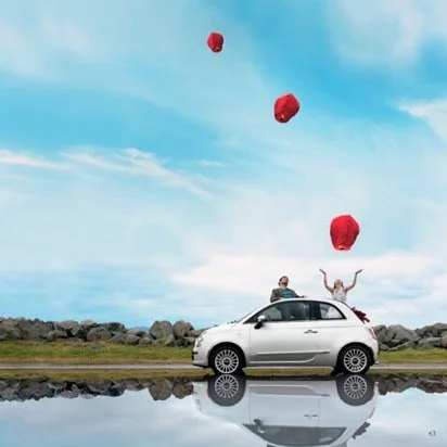 People throwing red lanterns into the air from a car parked near a rocky shoreline with a partly cloudy sky.