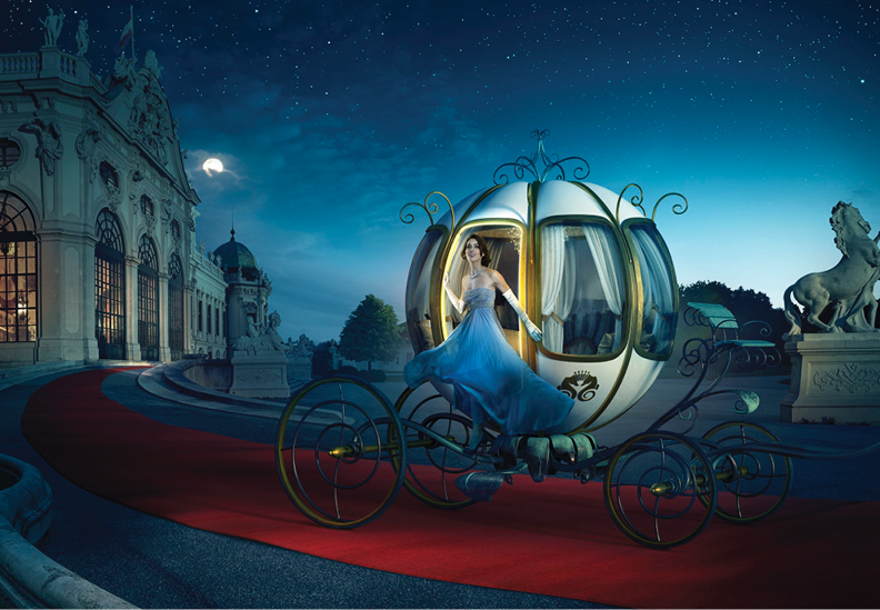 A woman in a blue gown sitting inside a glowing fairy-tale pumpkin carriage on a red carpet outside a grand palace at night, with a starry sky and moon in the background.