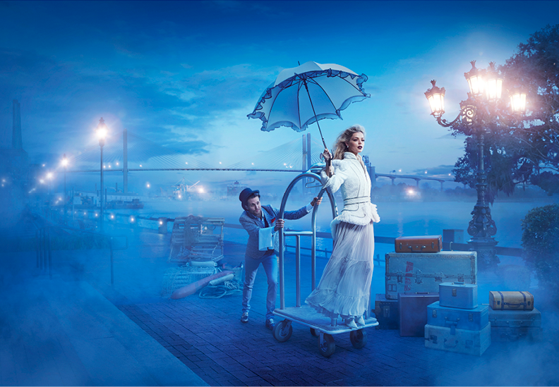 A woman in a white dress is standing on a moving cart holding an umbrella, while a man behind her pushes the cart. The scene is set on a foggy riverside promenade with streetlights illuminating the area.
