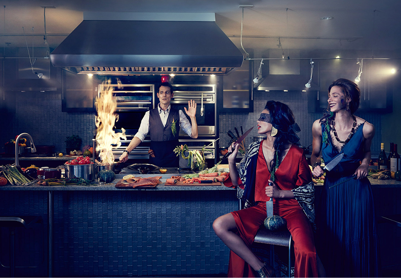Two women dressed in glamorous outfits, one with a blindfold and holding a knife, and the other smiling and holding a large knife, are in a modern kitchen with a chef in the background. The chef appears to be surprised as a pot on the stove catches f