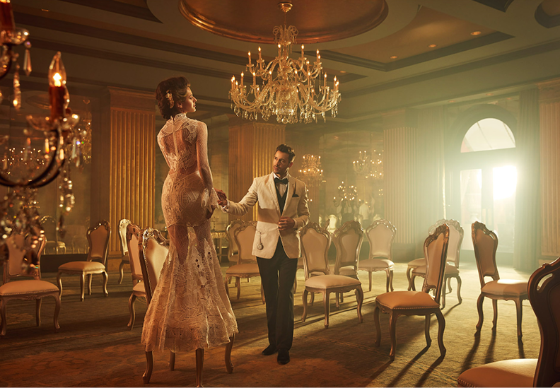 A man in a tuxedo and a woman in an elegant lace gown dancing in a luxurious ballroom with chandeliers and ornate chairs.