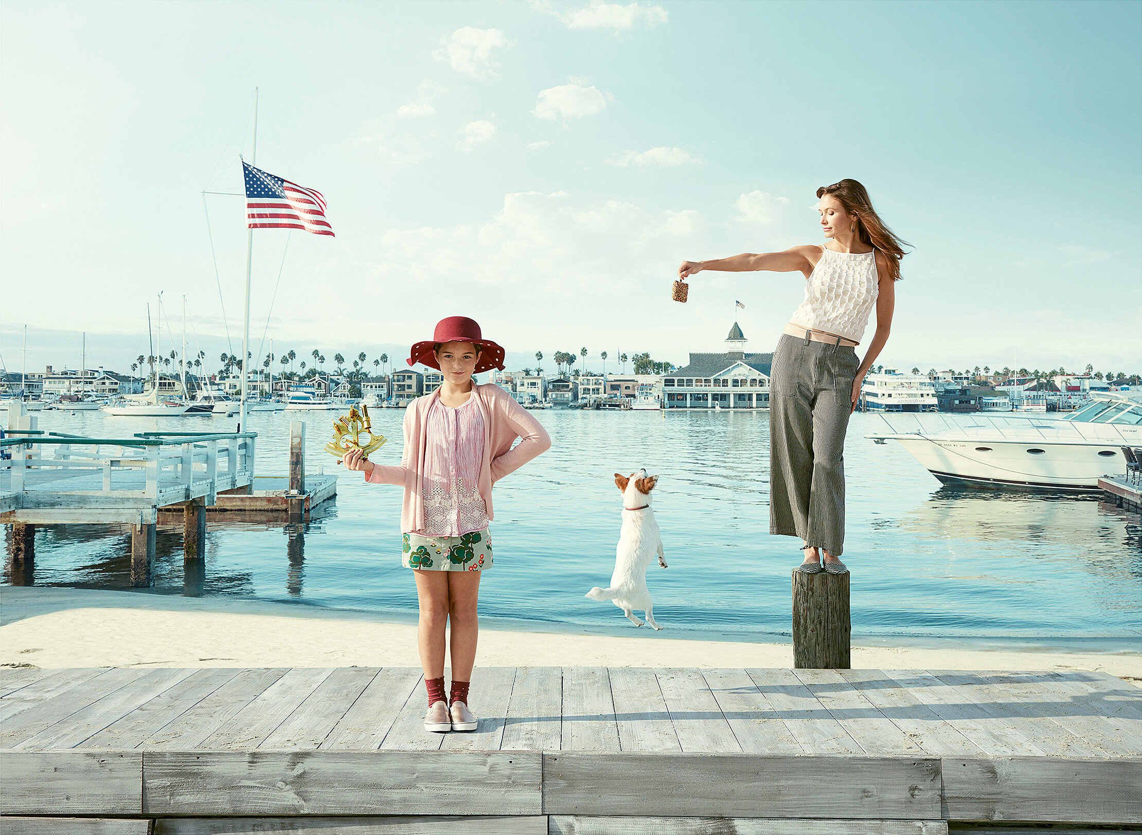 A woman and a girl with a dog near a marina with boats. The woman is balancing on a wooden post, handing a small object to the girl, who is holding a small sailboat model. The girl is wearing a large red hat, pink sweater, and green shorts. The woman