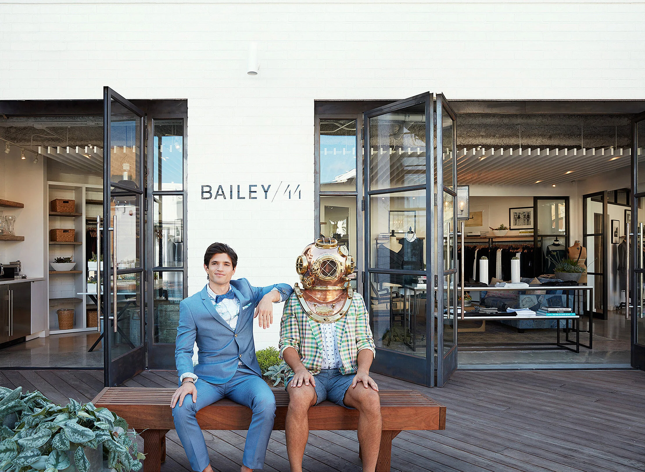 Two men sit on a wooden bench outside a modern boutique store with open glass doors. One man is dressed in a blue suit, and the other is wearing a vintage deep-sea diver helmet with casual clothing.