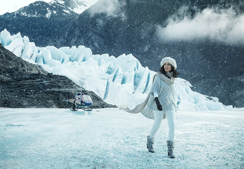 A woman dressed in winter clothing standing on ice near a glacier with a helicopter in the background.