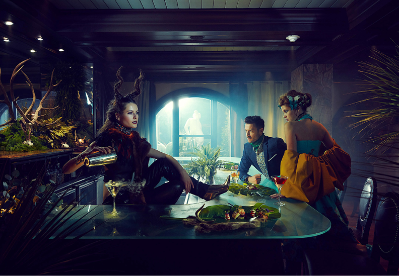 Three people dressed in elaborate costumes sitting around a dining table filled with food and drinks in a dimly lit room with dark wood paneling and large window.