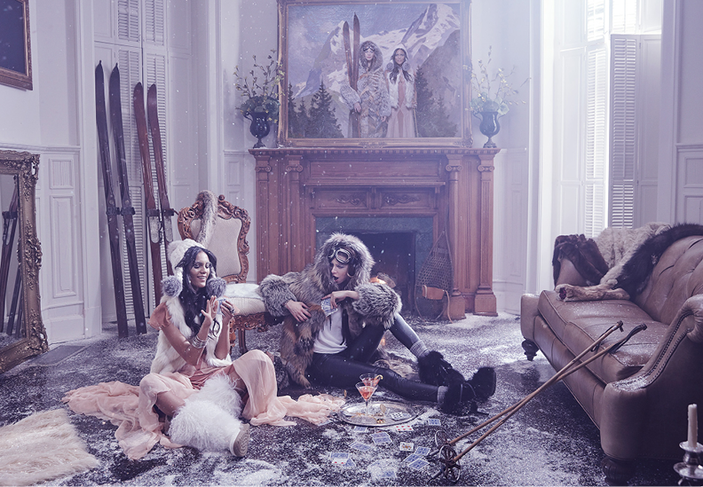 Two women dressed in winter clothing with faux fur coats and accessories sit in a disheveled living room with snow on the floor. One woman has glasses and is holding a drink, the other is sitting on the floor near a tray with drinks and playing cards