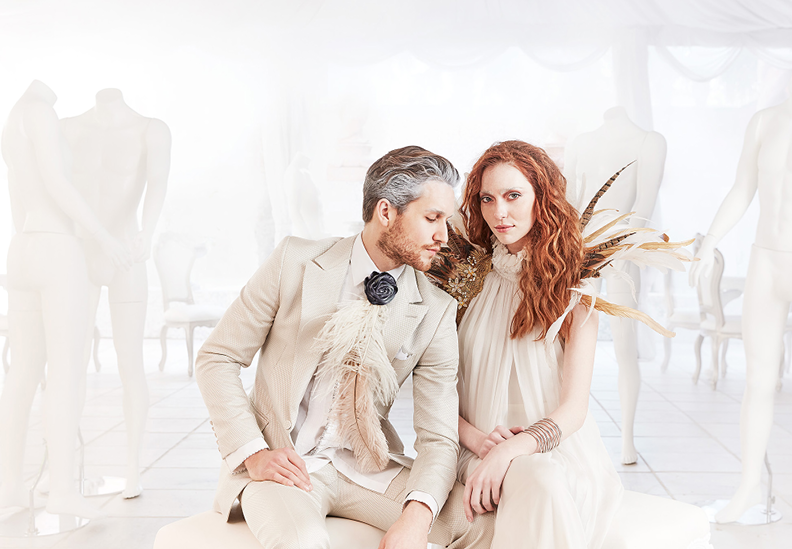 Man and woman sitting together in a bright, white room with mannequins, wearing elegant clothing with feather accents.
