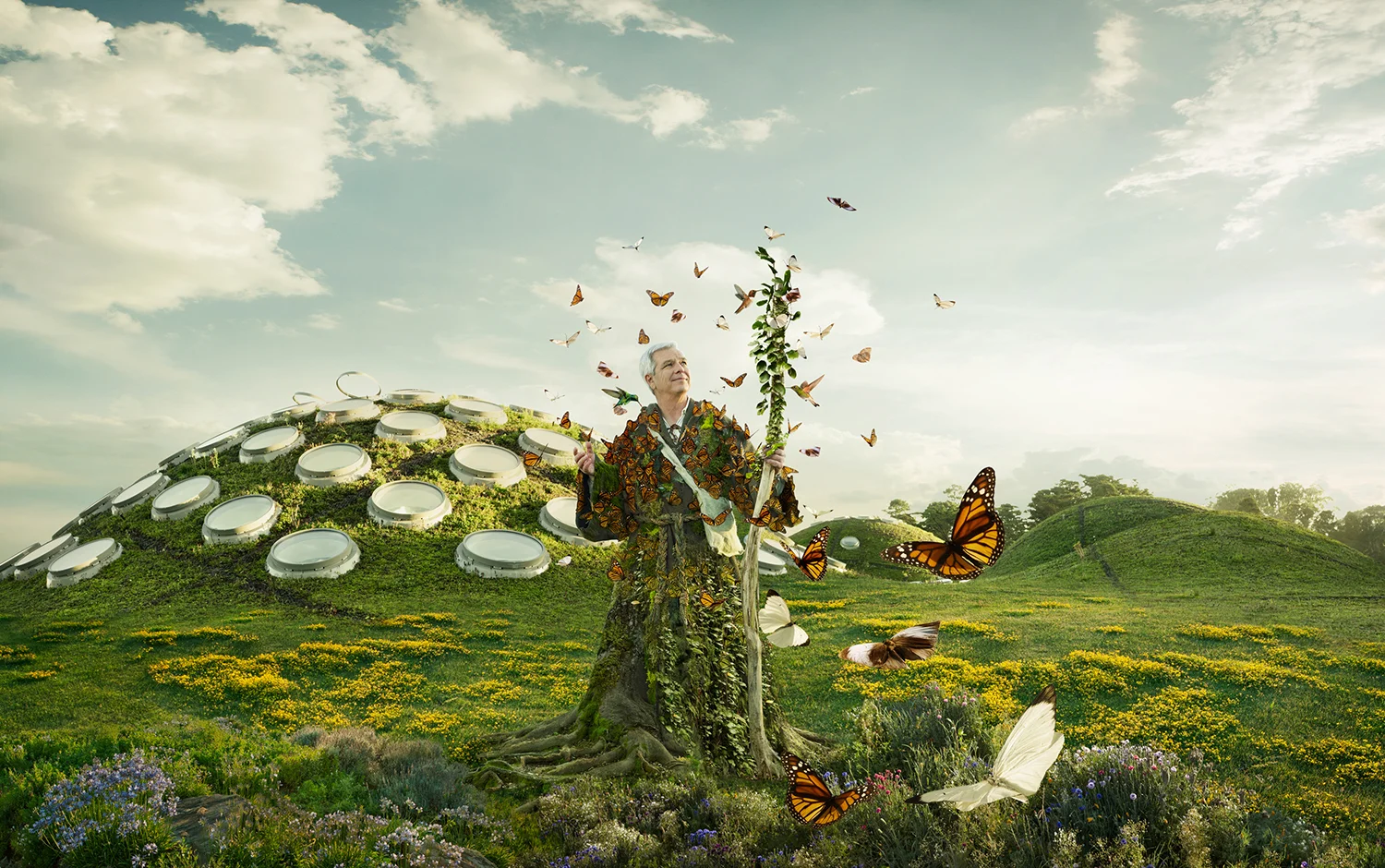 A man in a camouflage jacket stands next to a tree with butterflies flying around, in a lush green field with yellow flowers and hills in the background.