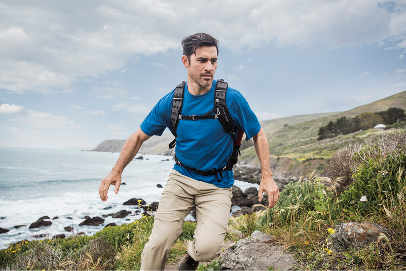A man hiking on a coastal trail with rocks, ocean, and grassy hills in the background.