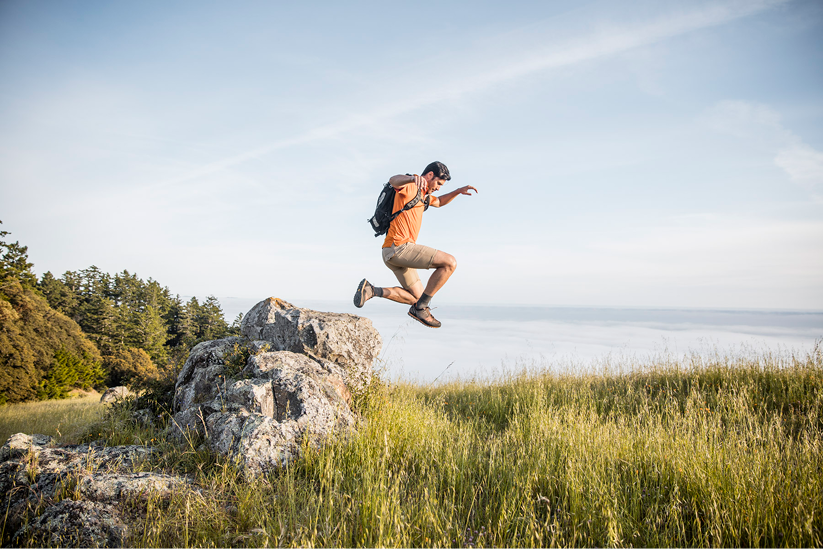 A man wearing an orange shirt and beige shorts jumping over large rocks in a grassy field beside the ocean on a clear day.