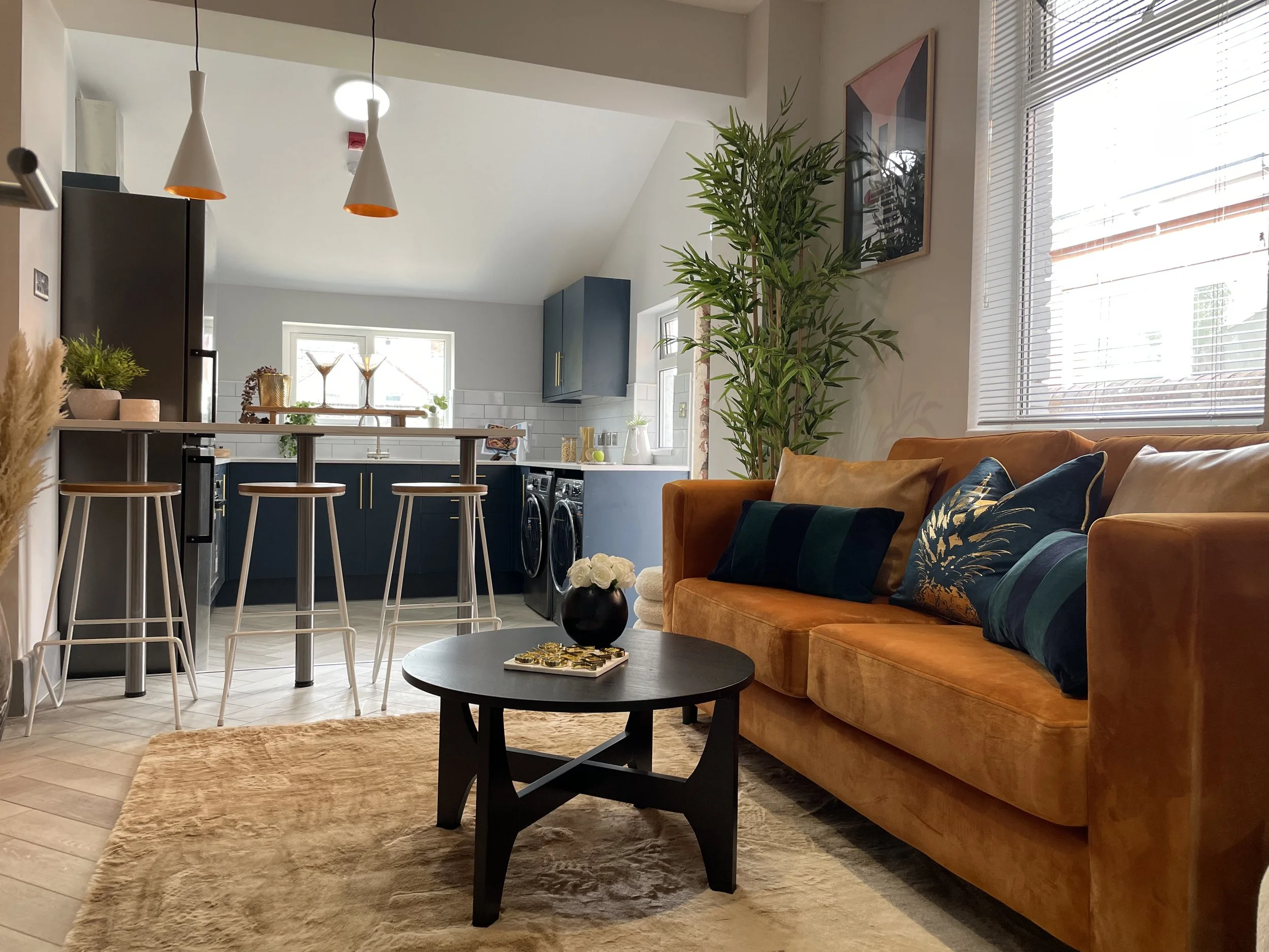 Living room with a tan sofa, decorative pillows, a black coffee table with a vase of white roses, a beige area rug, and large windows with blinds. The kitchen in the background has dark blue cabinets, a white backsplash, a window, and bar stools at a counter.