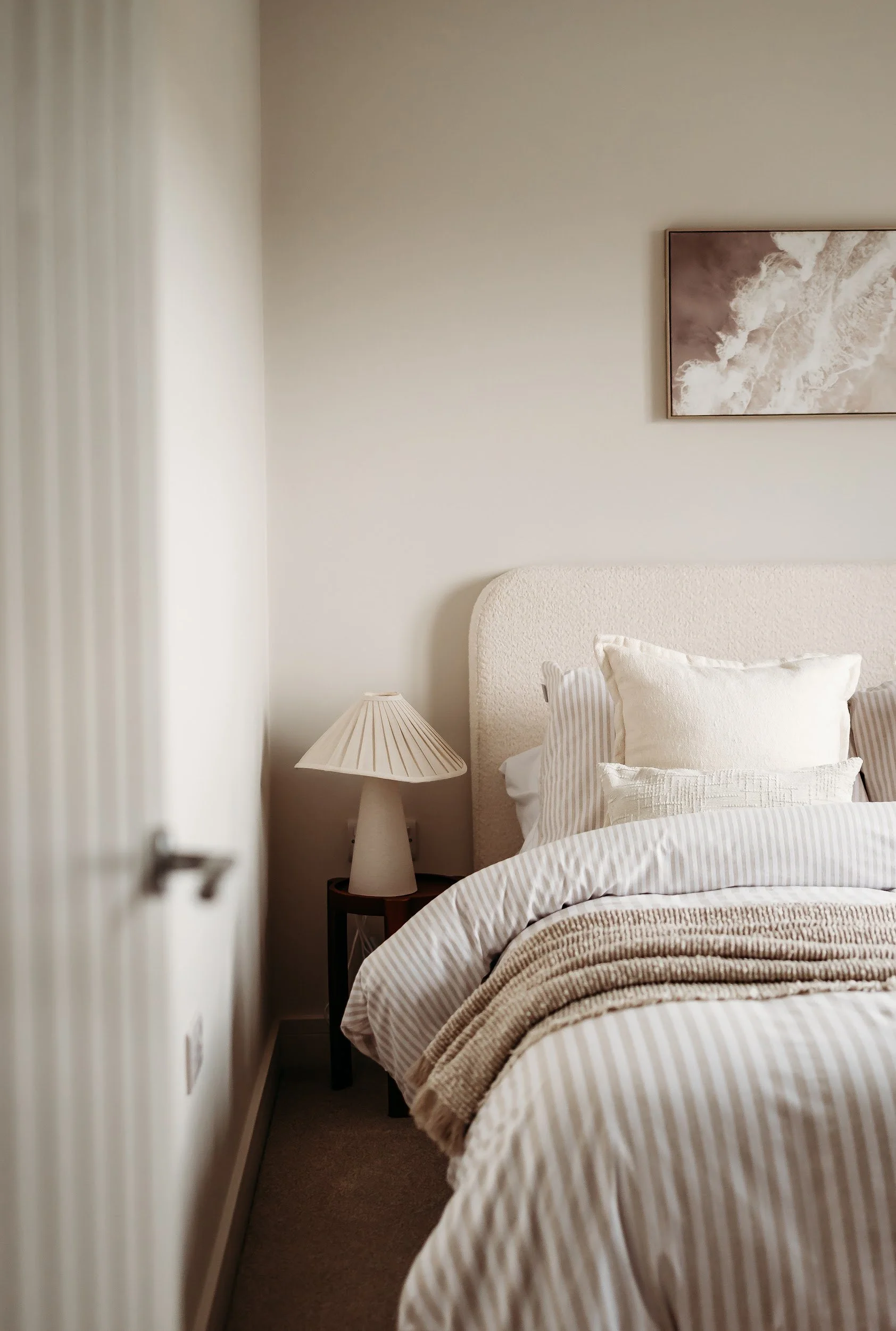 A neatly made bed with multiple pillows and a brown faux fur blanket in a modern bedroom, with a textured beige headboard, a white bedside table with a lamp and tray, and an abstract gold and gray painting on a white wall.