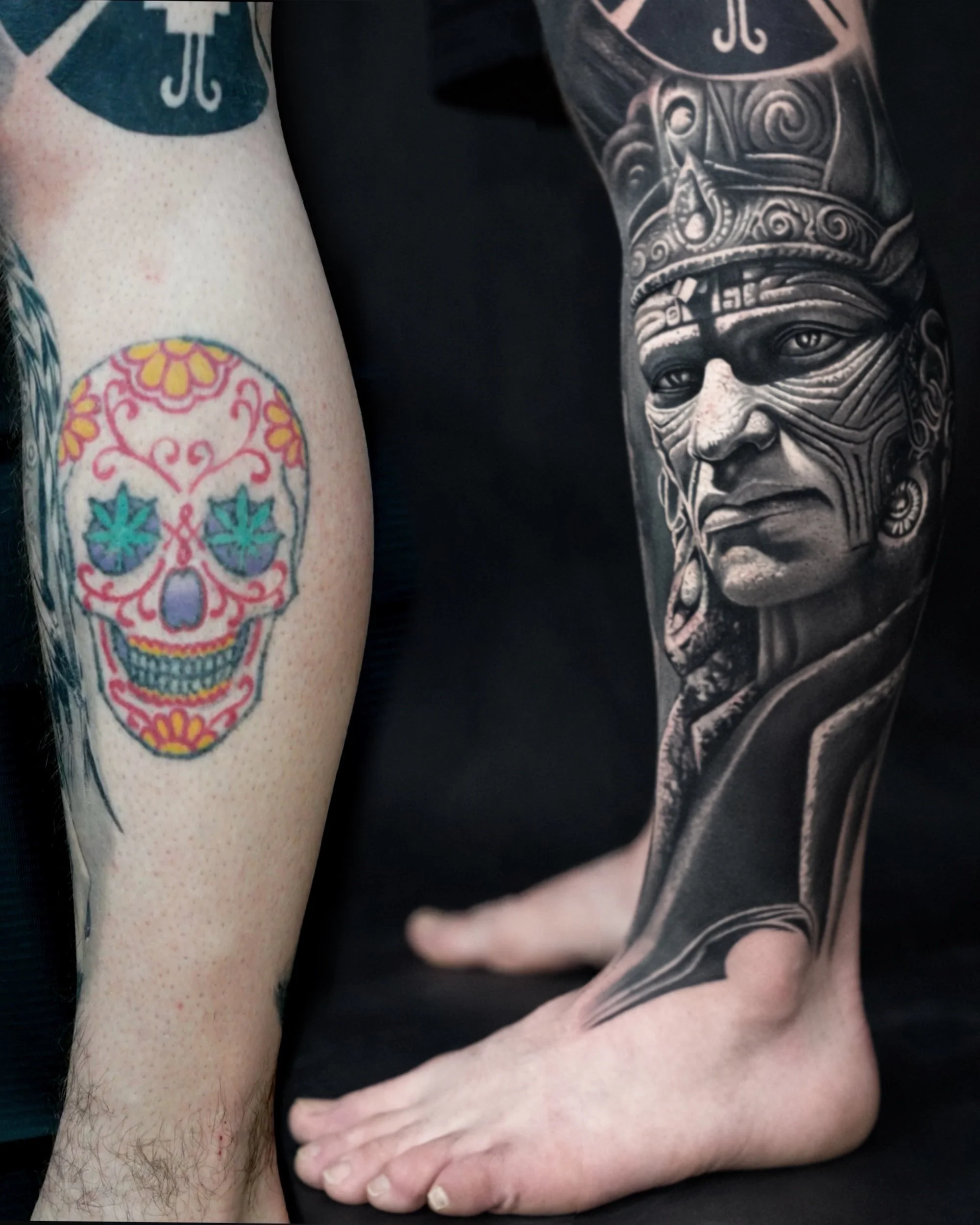 Das Bild zeigt zwei Tattoos auf den Armen und Beinen einer Person. Der eine Arm hat ein farbenfrohes Totenkopf-Tattoo im Day of the Dead Stil, und das andere Bein hat ein realistisches schwarze und graues Tribal- oder Mythologietattoo eines menschlic