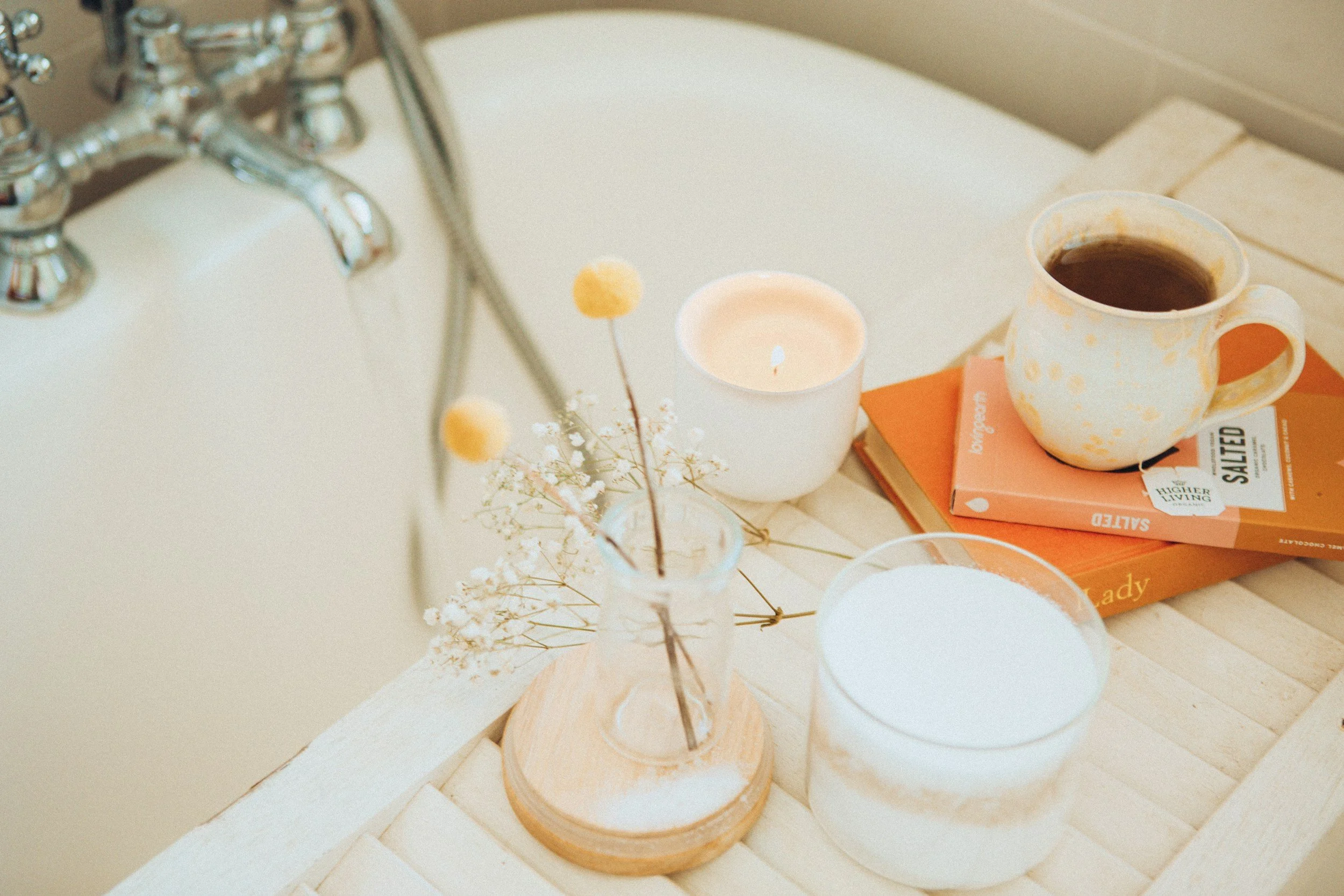 A cozy bathroom scene with a bathtub, a lit candle, a book, a cup of coffee, and a vase with dried flowers on a wooden table.