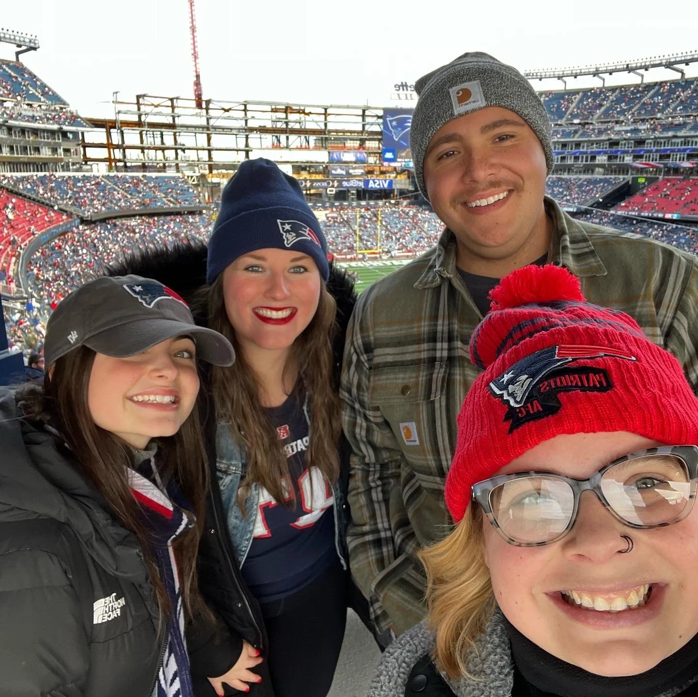 Four people at a football game in a stadium, each wearing New England Patriots apparel and hats, smiling for a selfie with the stadium seats and field in the background.