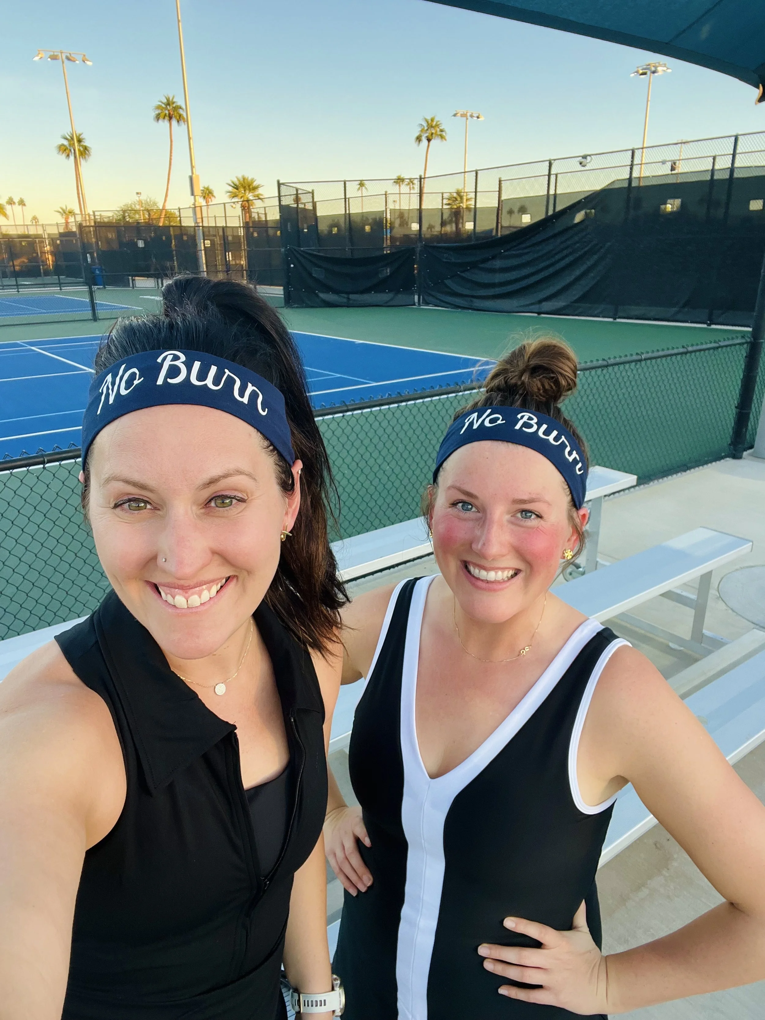 Two women wearing black and white sports outfits and blue headbands with 'No Bump' written on them, taking a selfie at an outdoor tennis court. The court has palm trees and lighting poles in the background.