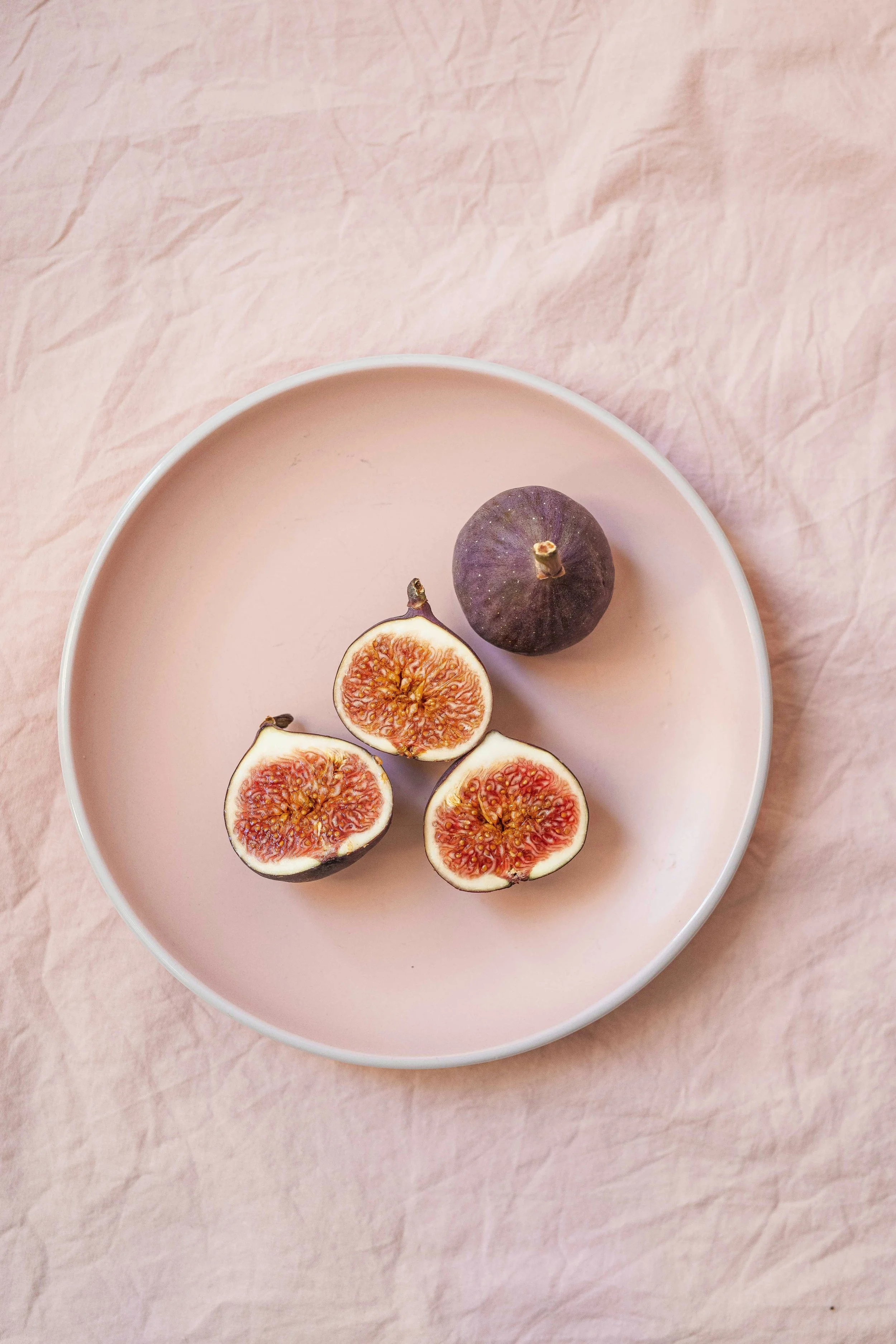 A pink plate with three halved figs and one whole fig on a wrinkled pink fabric background.