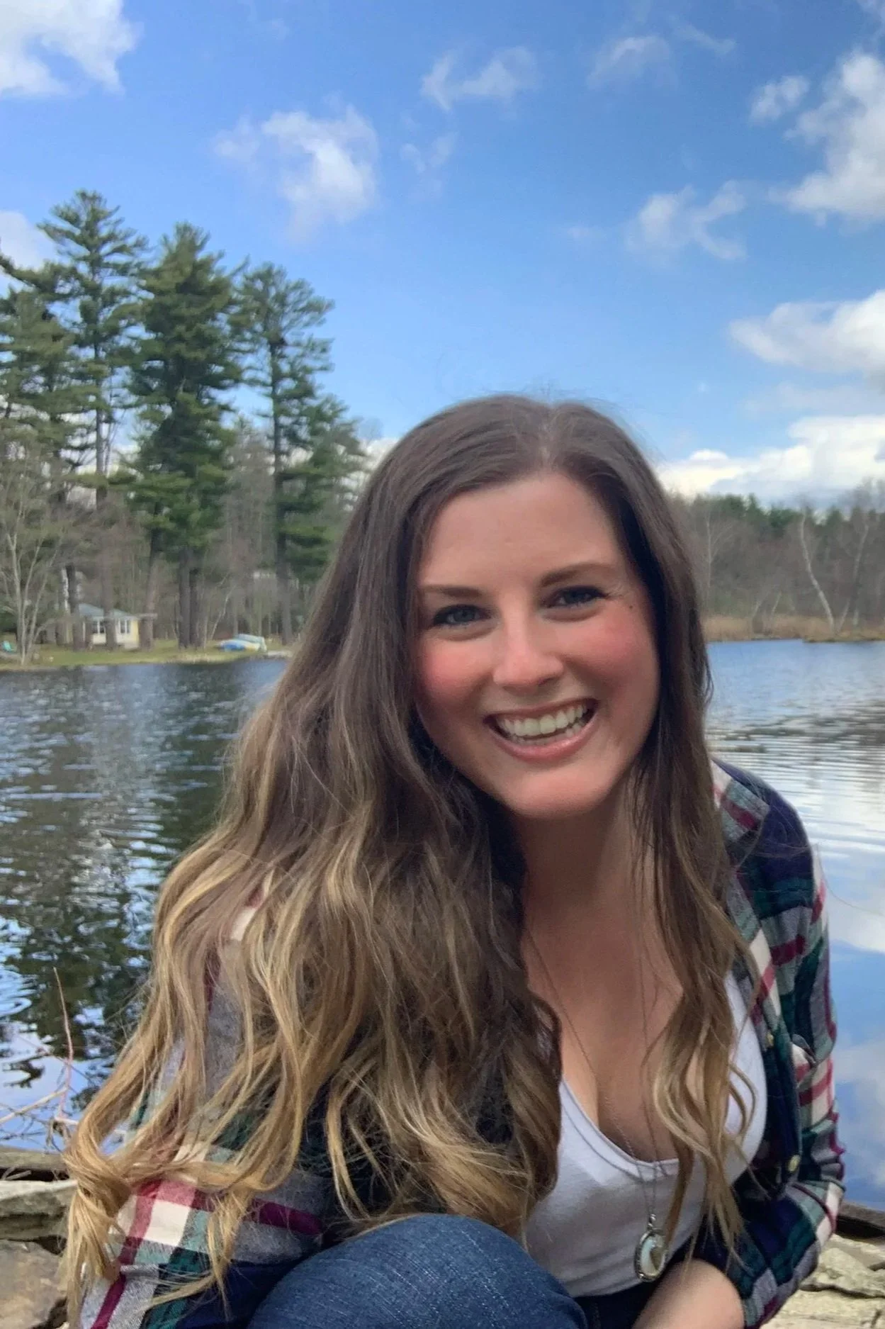 A young woman with long wavy hair smiling while sitting outdoors beside a lake, with trees and houses in the background and a blue sky with some clouds.