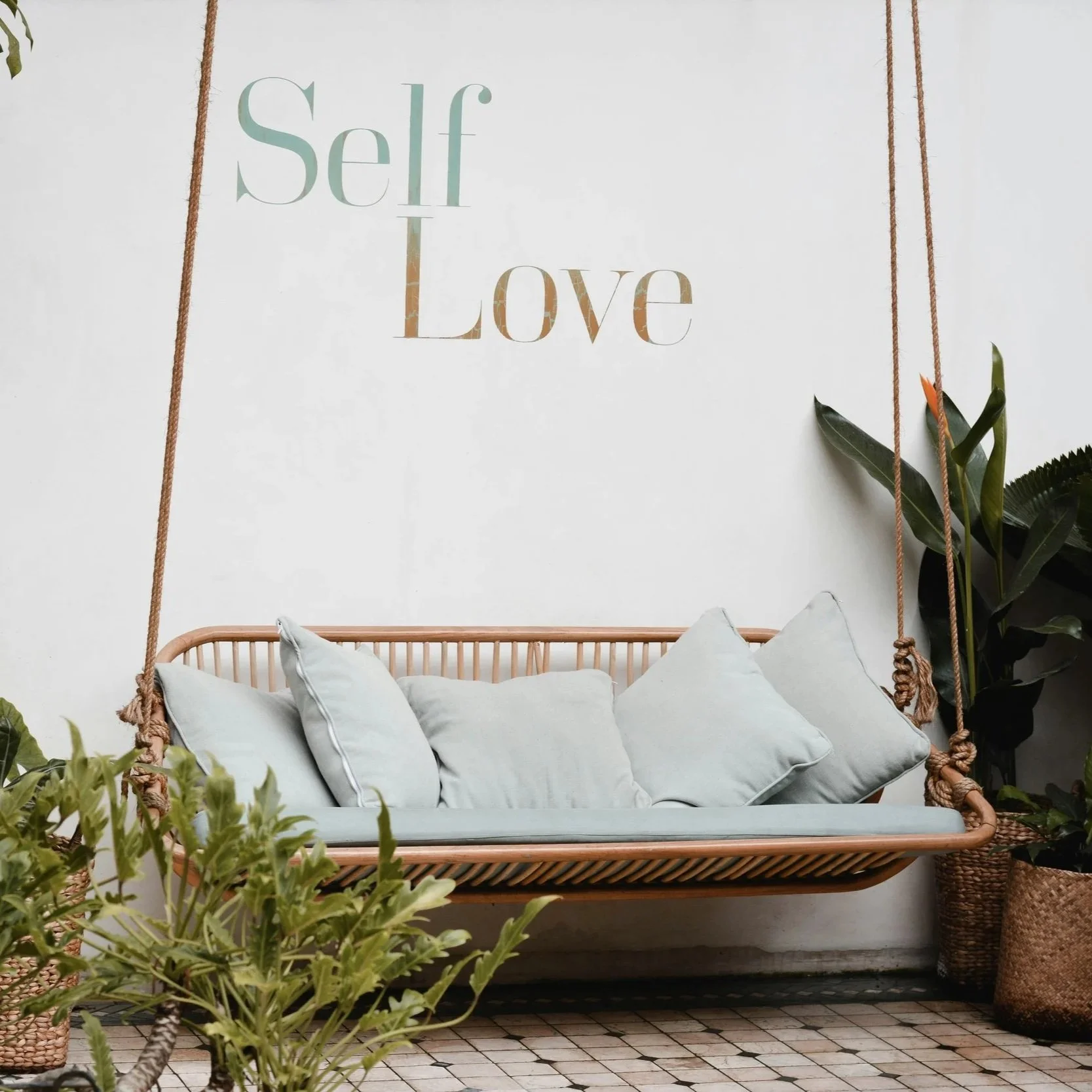 A cozy indoor seating area with a rattan hanging bench filled with white pillows, surrounded by green plants, and a wall decoration with the words 'Self Love'.