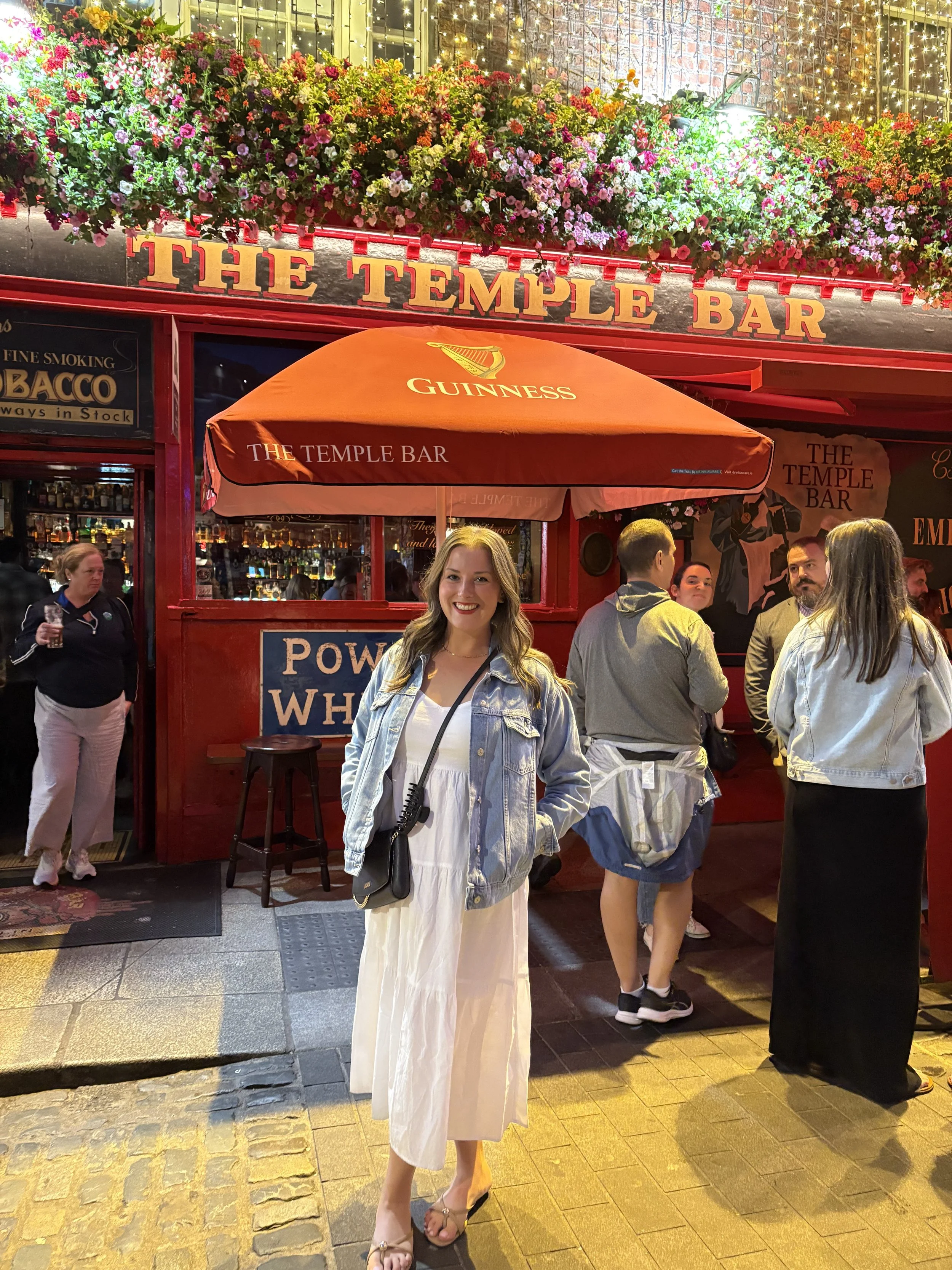 A woman standing outside a lively bar called The Temple Bar at night, with a bright red exterior, a Guinness-branded umbrella, and people socializing inside and outside.