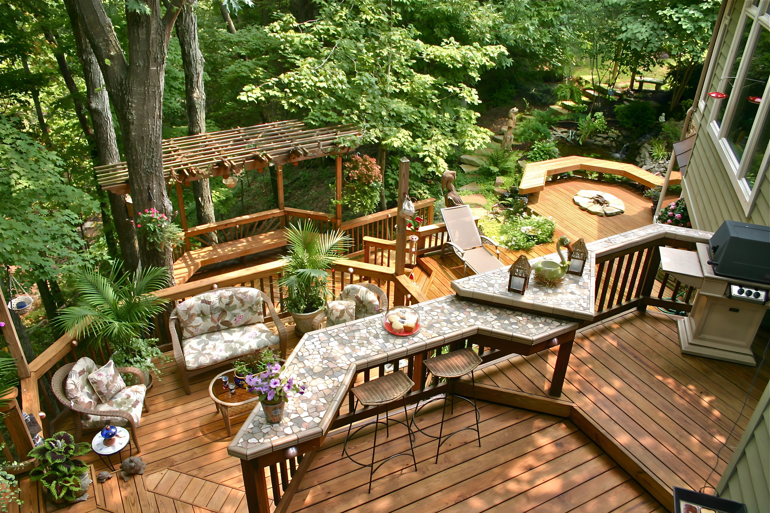A multi-level wooden deck with outdoor furniture, potted plants, a barbecue, and surrounded by lush green trees and garden decorations.