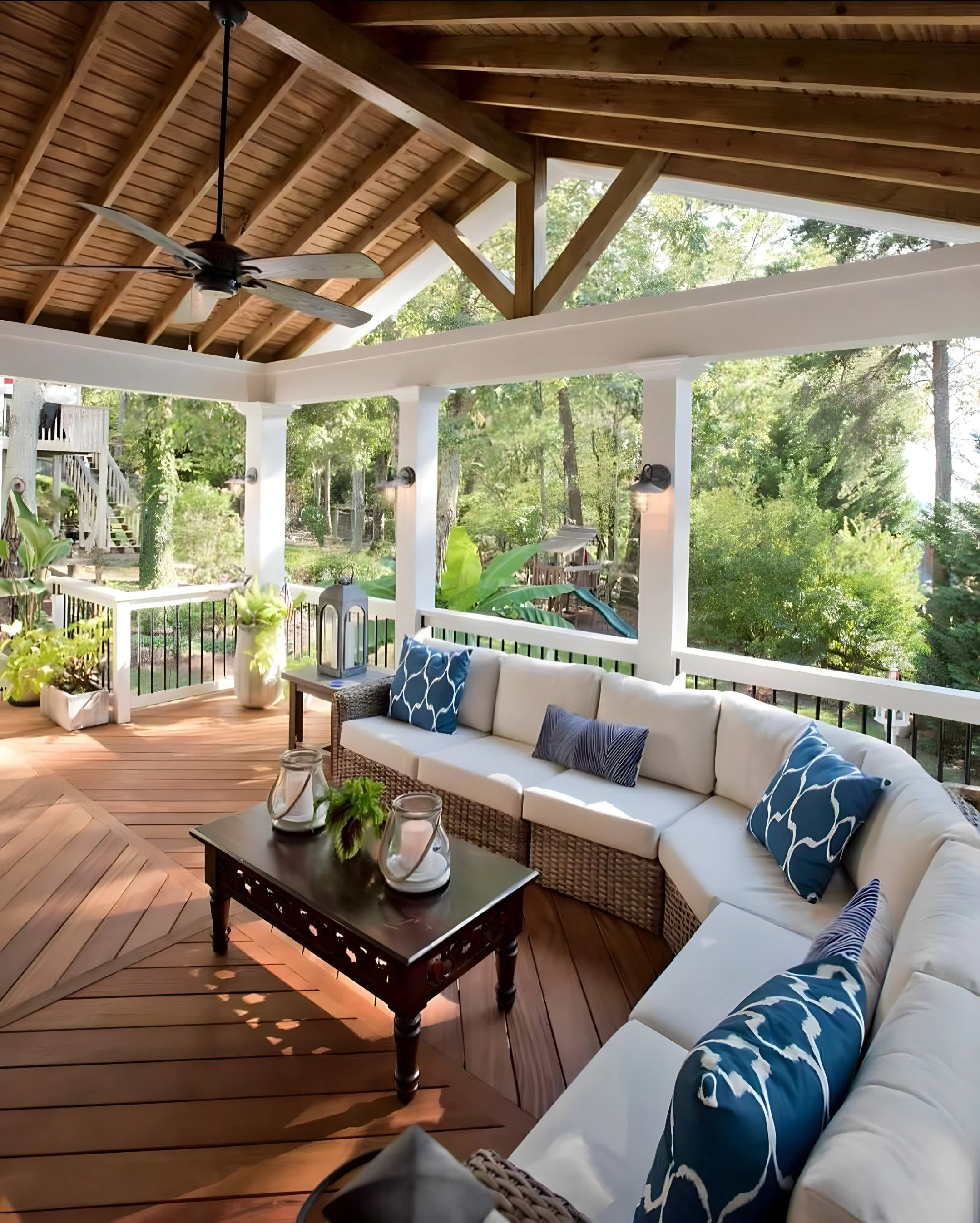 Covered outdoor porch with white corner sofa with blue patterned cushions, a wooden coffee table with lanterns and decor, potted plants, ceiling fan, and lush green trees in the background.