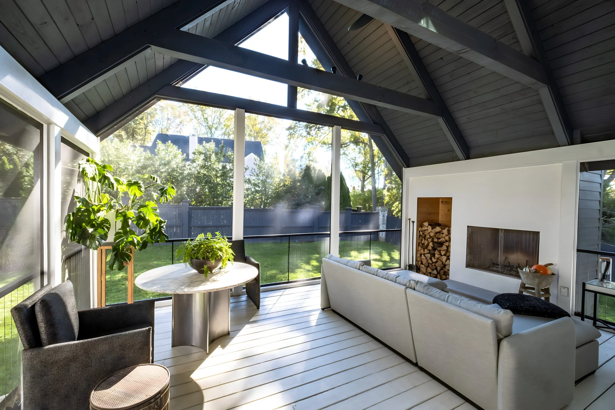 Sunlit screened porch with white and black furniture, a wood-burning fire fireplace, wood storage, potted plants, and large glass windows facing a green backyard.