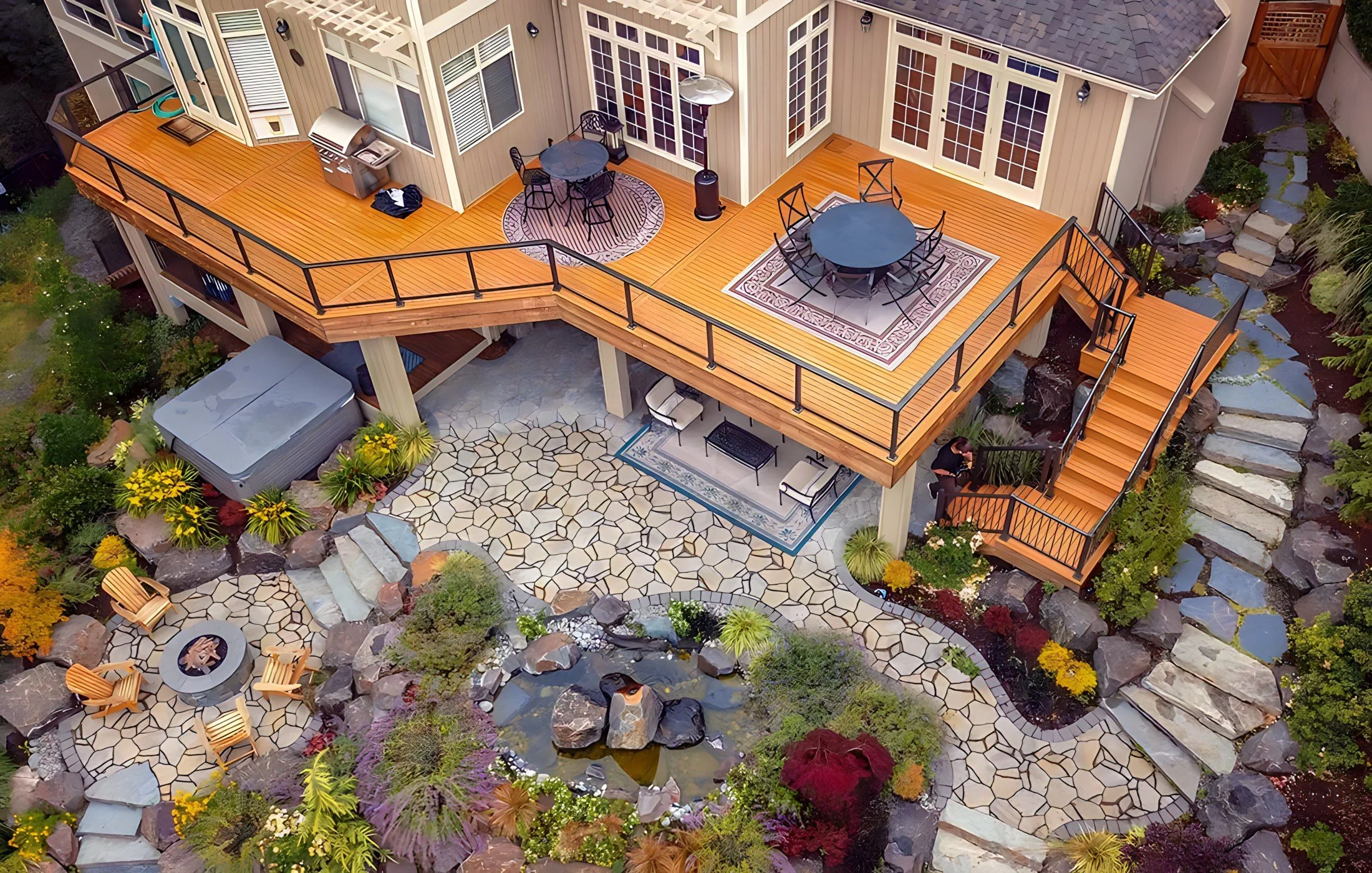 Aerial view of a two-story house with a spacious wooden deck, outdoor furniture, a hot tub, and landscaped garden with a pond, stones, and plants.