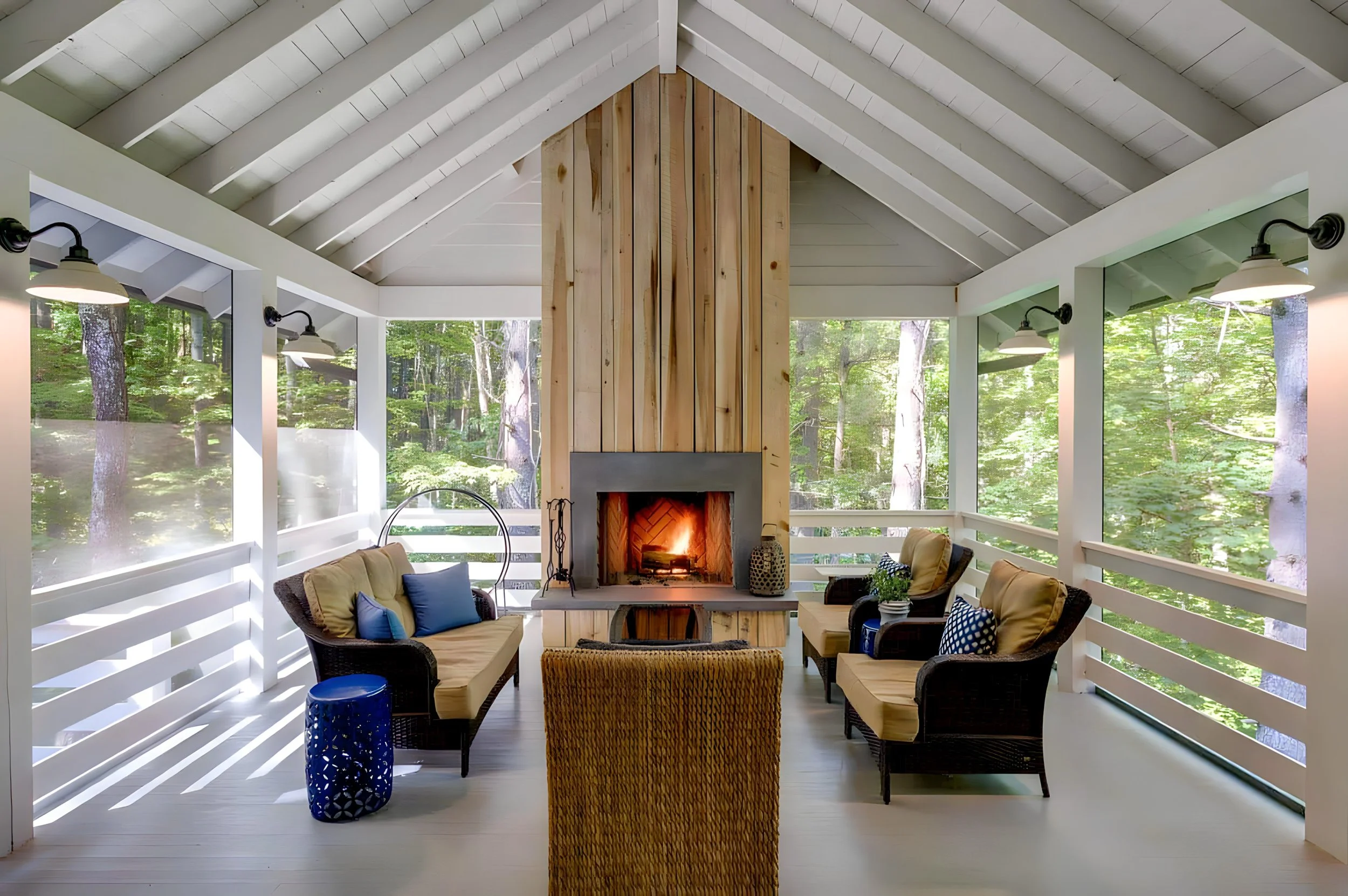 Enclosed porch with a fireplace, wicker chairs, and outdoor cushions surrounded by trees.