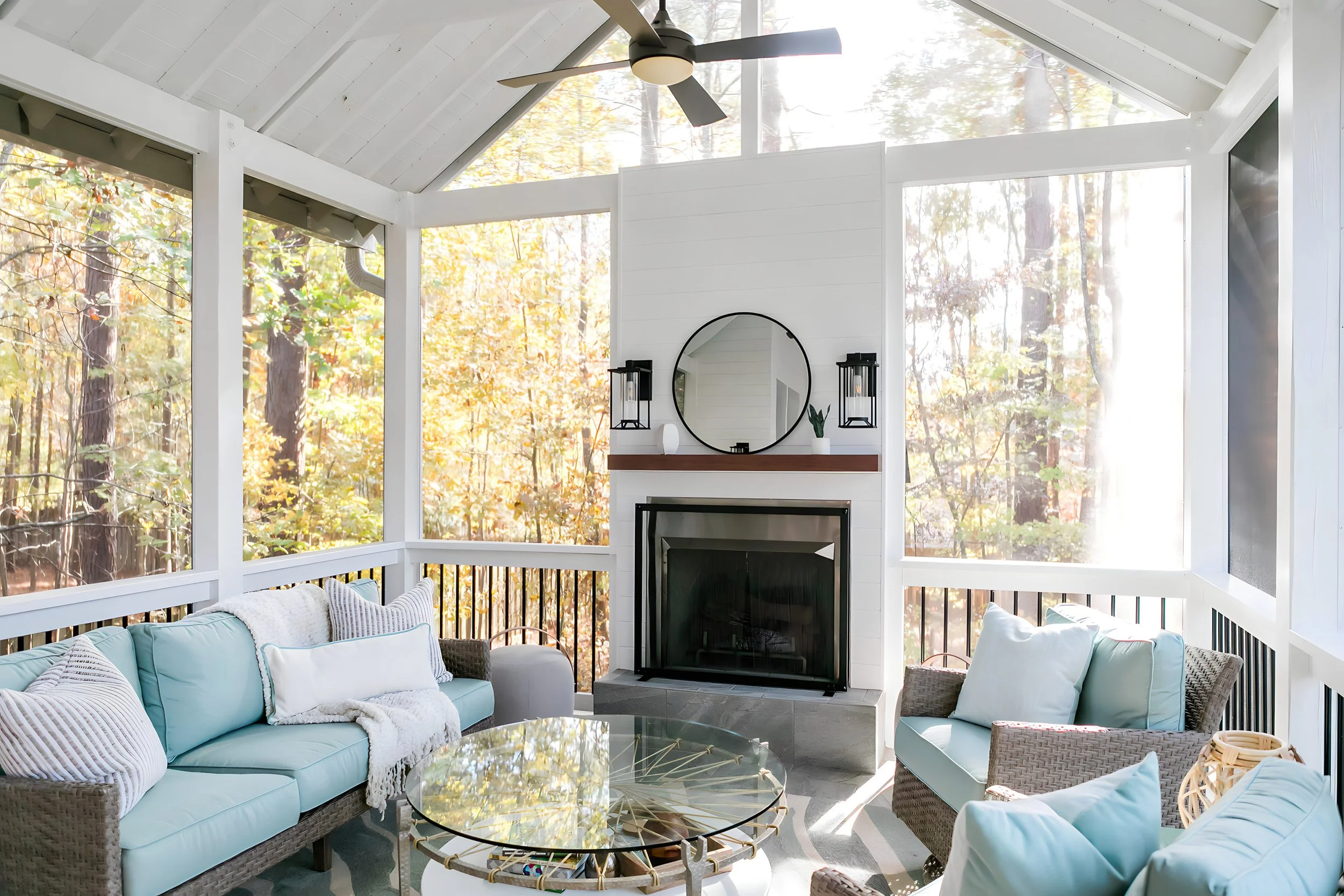 Sunlit screened porch with wicker furniture with light blue cushions and striped pillows, a glass coffee table, and a white fireplace with a black framed glass door, round mirror, and black lantern-style wall sconces, surrounded by oak trees with fall foliage.