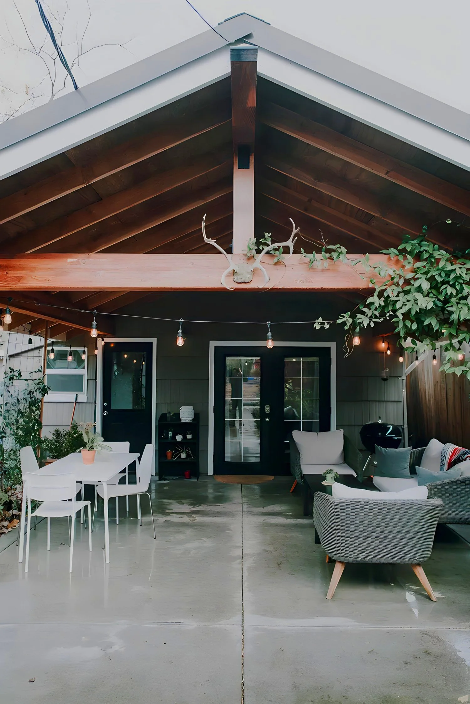 Covered outdoor patio with a dining table and chairs on the left, a seating area with armchairs and a sofa on the right, string lights hanging above, a decorative antler mounted on the wooden beam, and plants on the patio.