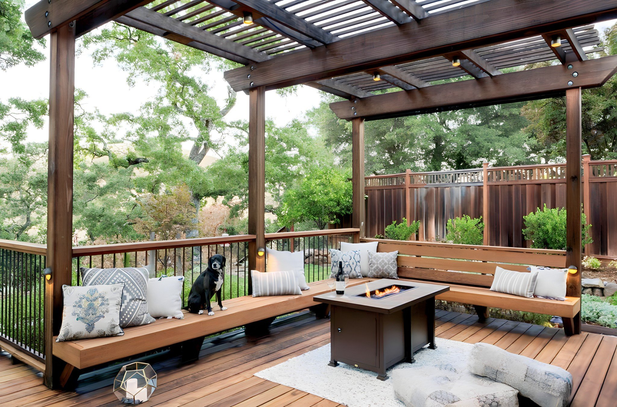 Outdoor patio with wooden deck, built-in bench with pillows, a fire pit table, and a dog sitting on the bench, surrounded by trees and a wooden fence.