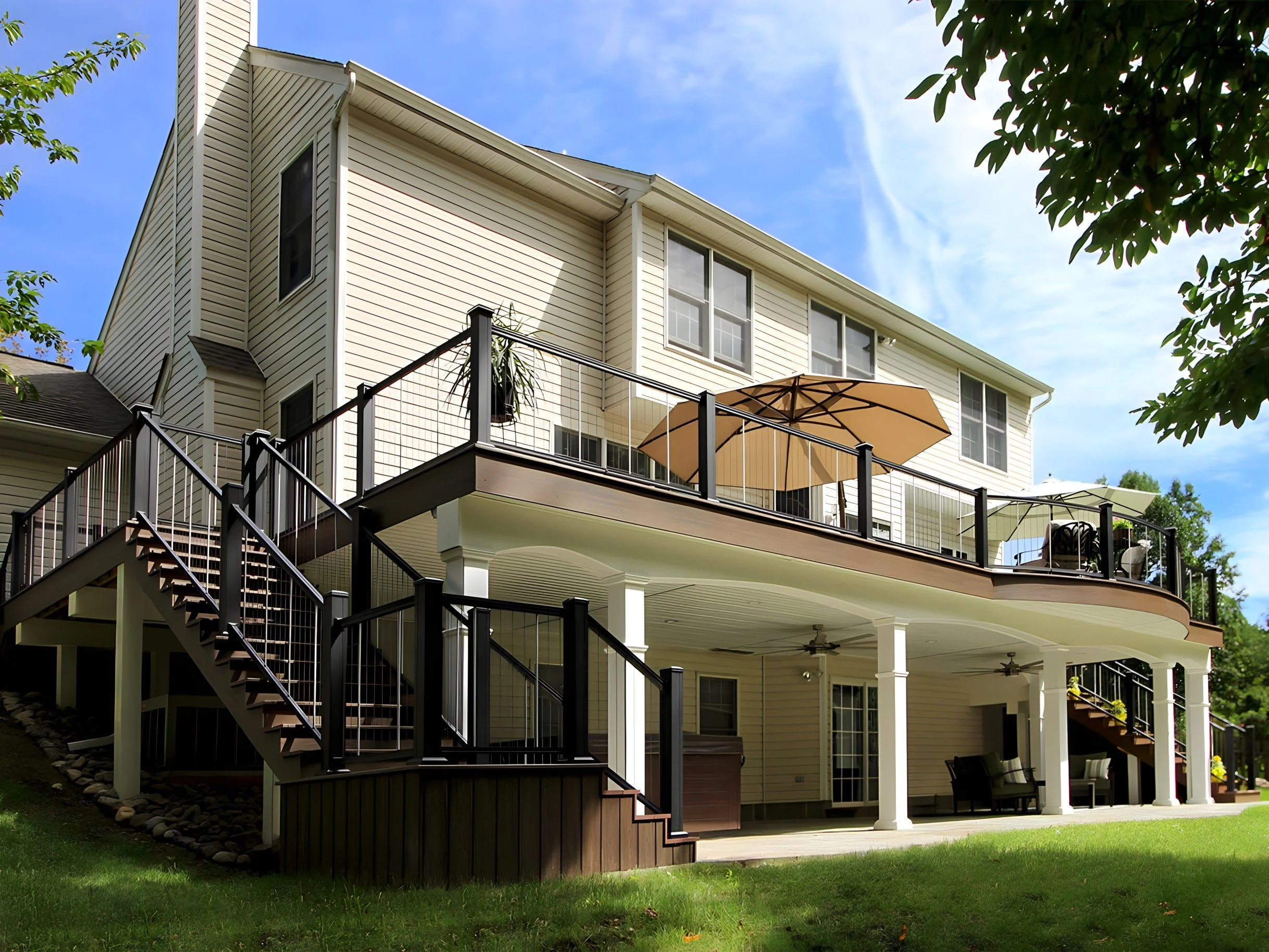Back view of a multi-story house with a large elevated deck, patio furniture, a large umbrella, and an outdoor setup, with a well-maintained lawn and trees in the surroundings.
