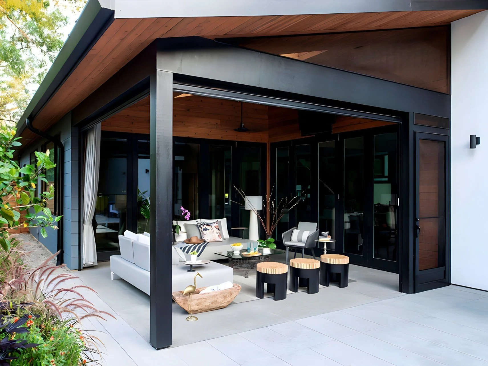 Modern outdoor patio with white seating, wooden stools, potted plants, and glass sliding doors leading into a house.