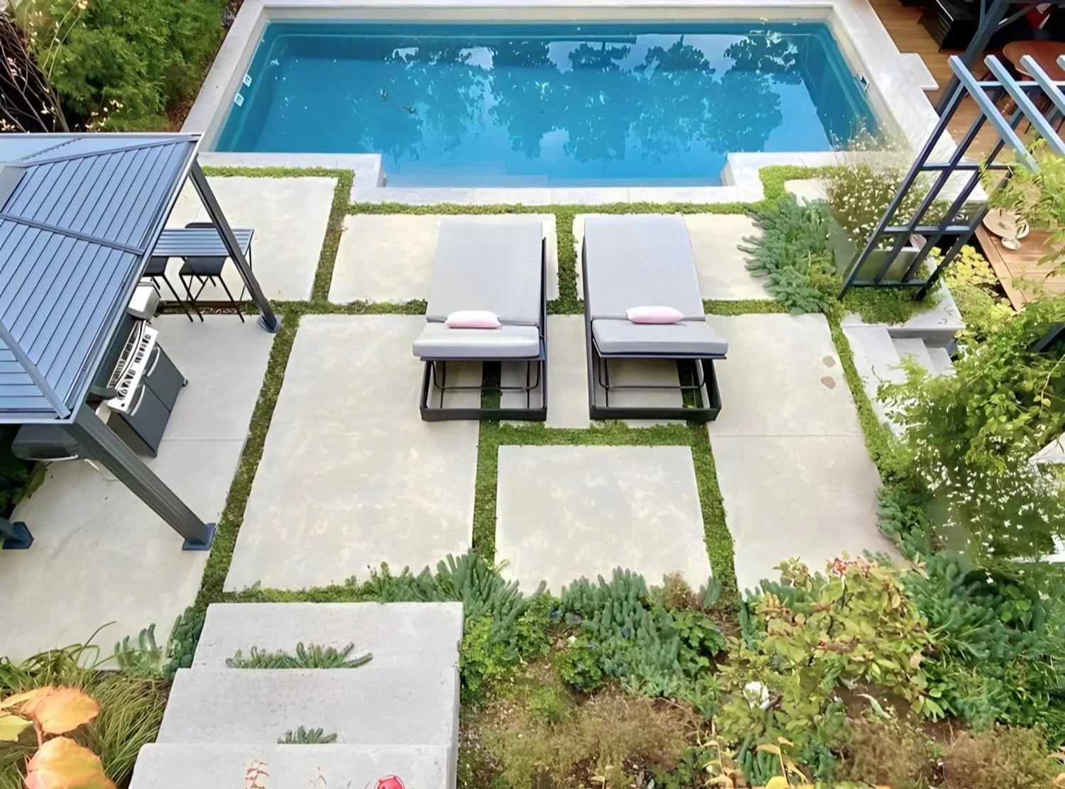 A backyard with a swimming pool, two lounge chairs with towels, a covered outdoor kitchen with a grill, and surrounding greenery.