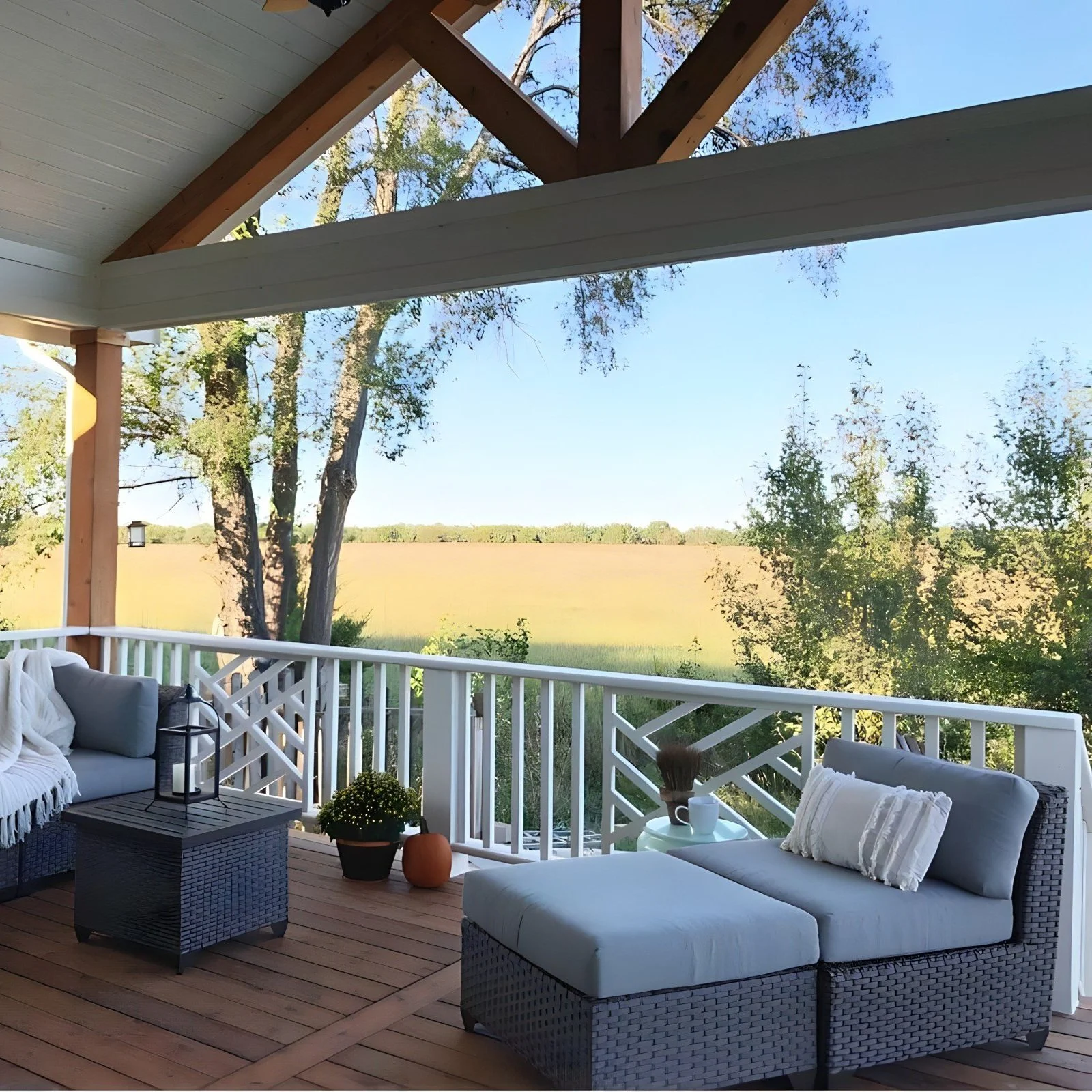 Covered porch with outdoor furniture including a cushioned lounge chair, a small table with a lantern, a potted plant, and a pumpkin, overlooking a field and trees under a blue sky.