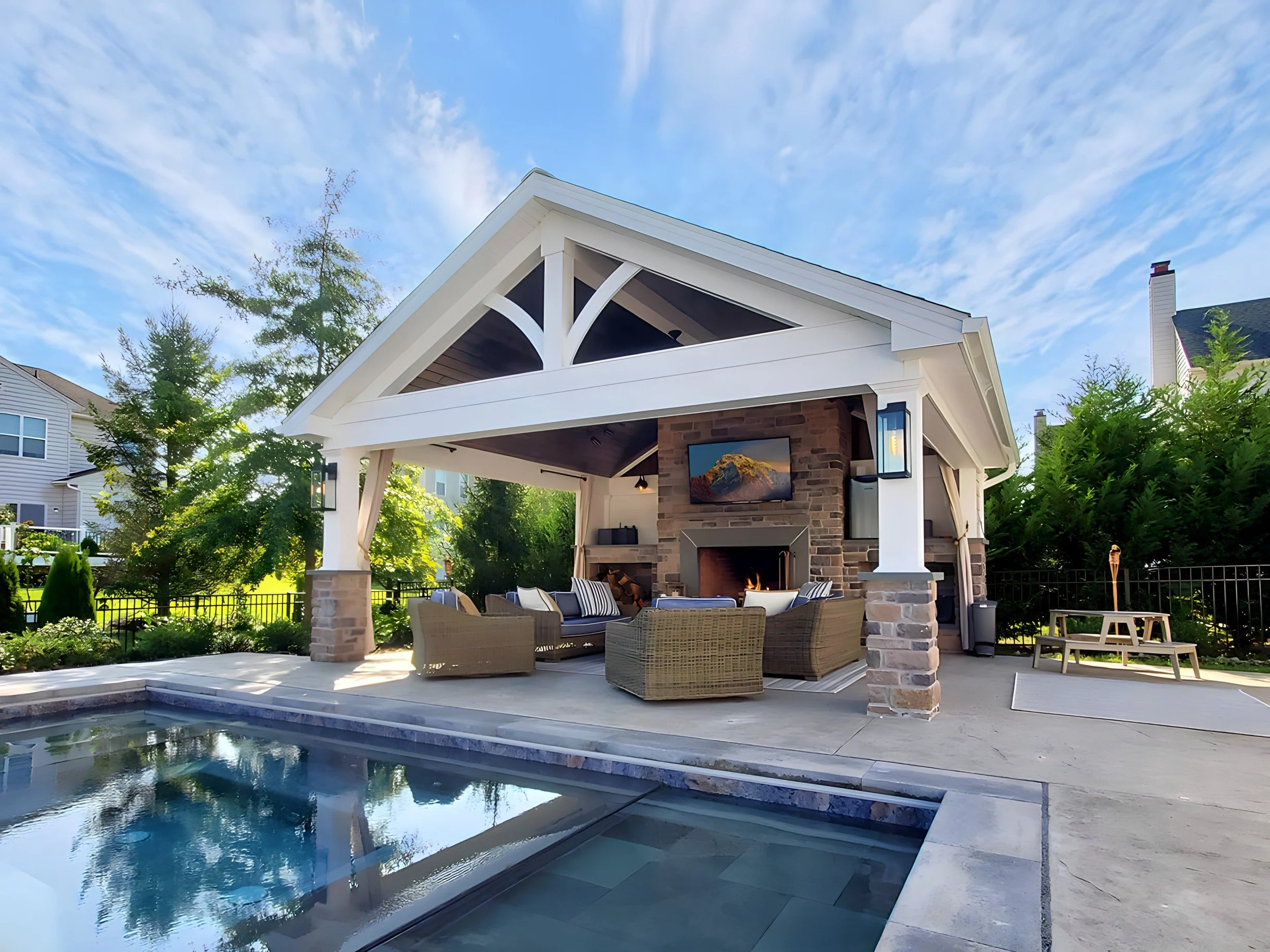 Modern backyard pool area with an open-air pavilion featuring a fireplace and television, surrounded by lush greenery and neighboring houses.
