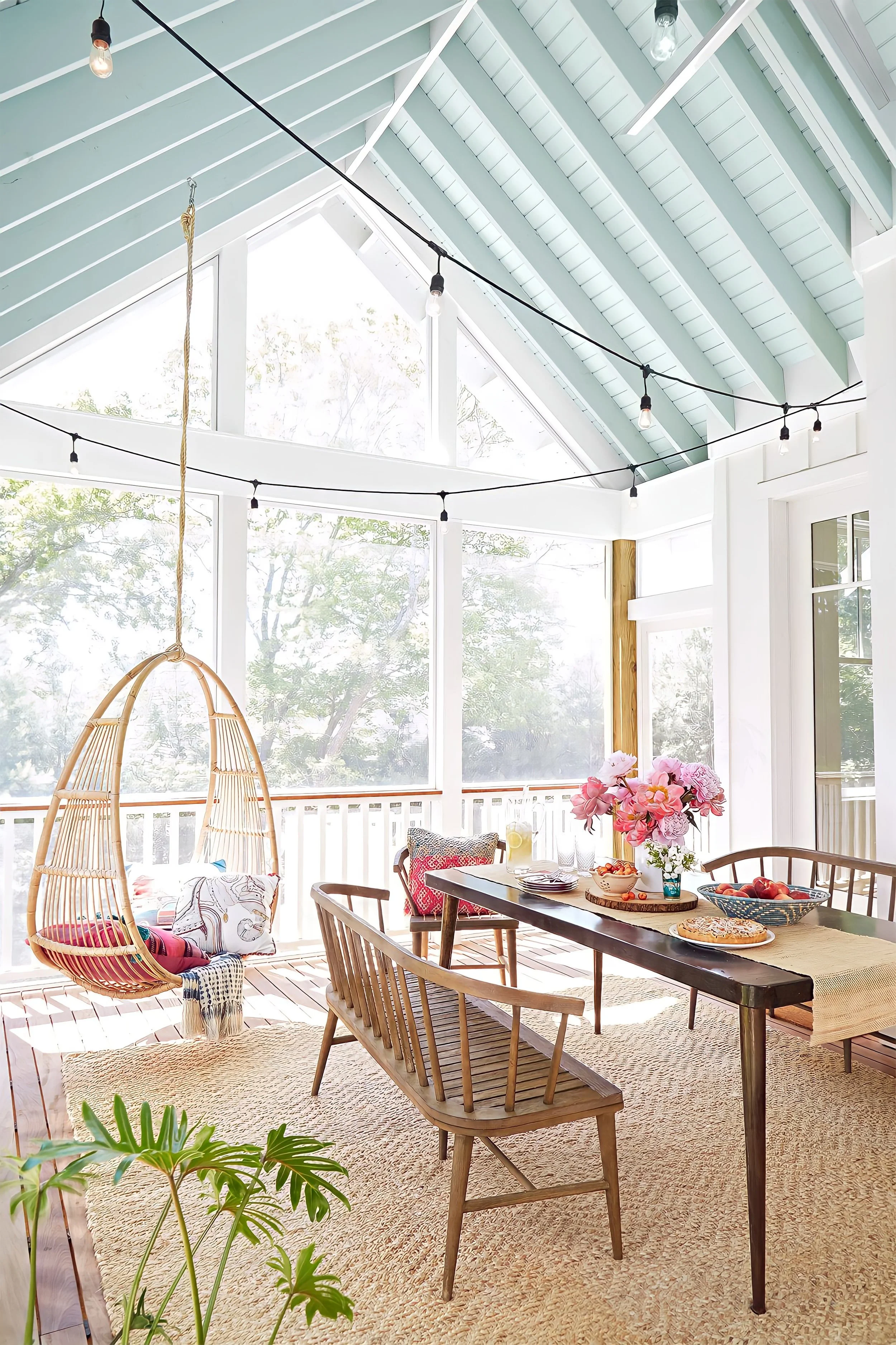 Bright sunroom with large windows, wooden dining table with tableware and flowers, hanging wicker chair with cushions, string lights, potted plant, and a light-colored rug.