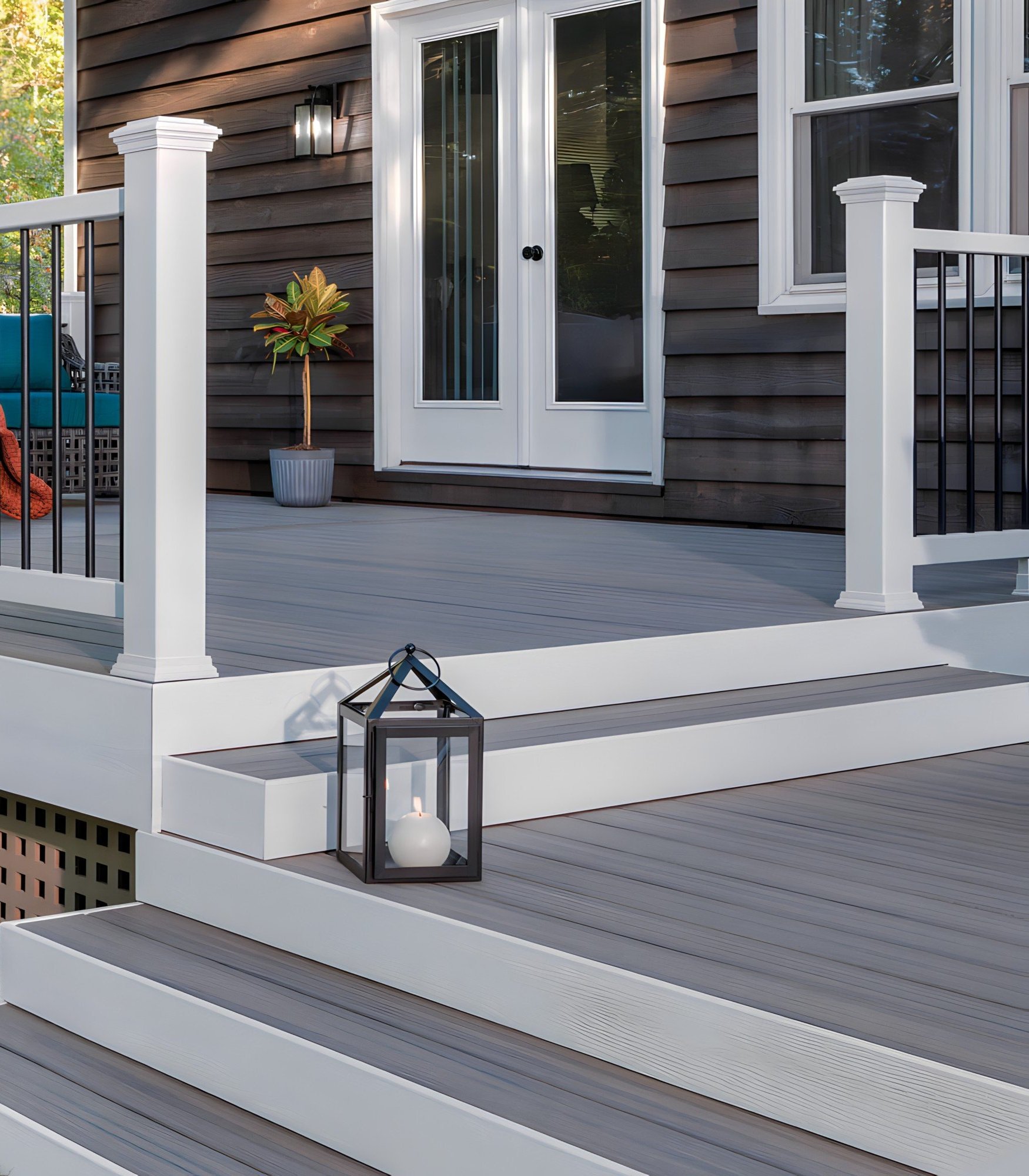 A modern outdoor deck with white steps, black metal lantern with a candle, potted plant, and glass door leading inside a house with dark wood siding.