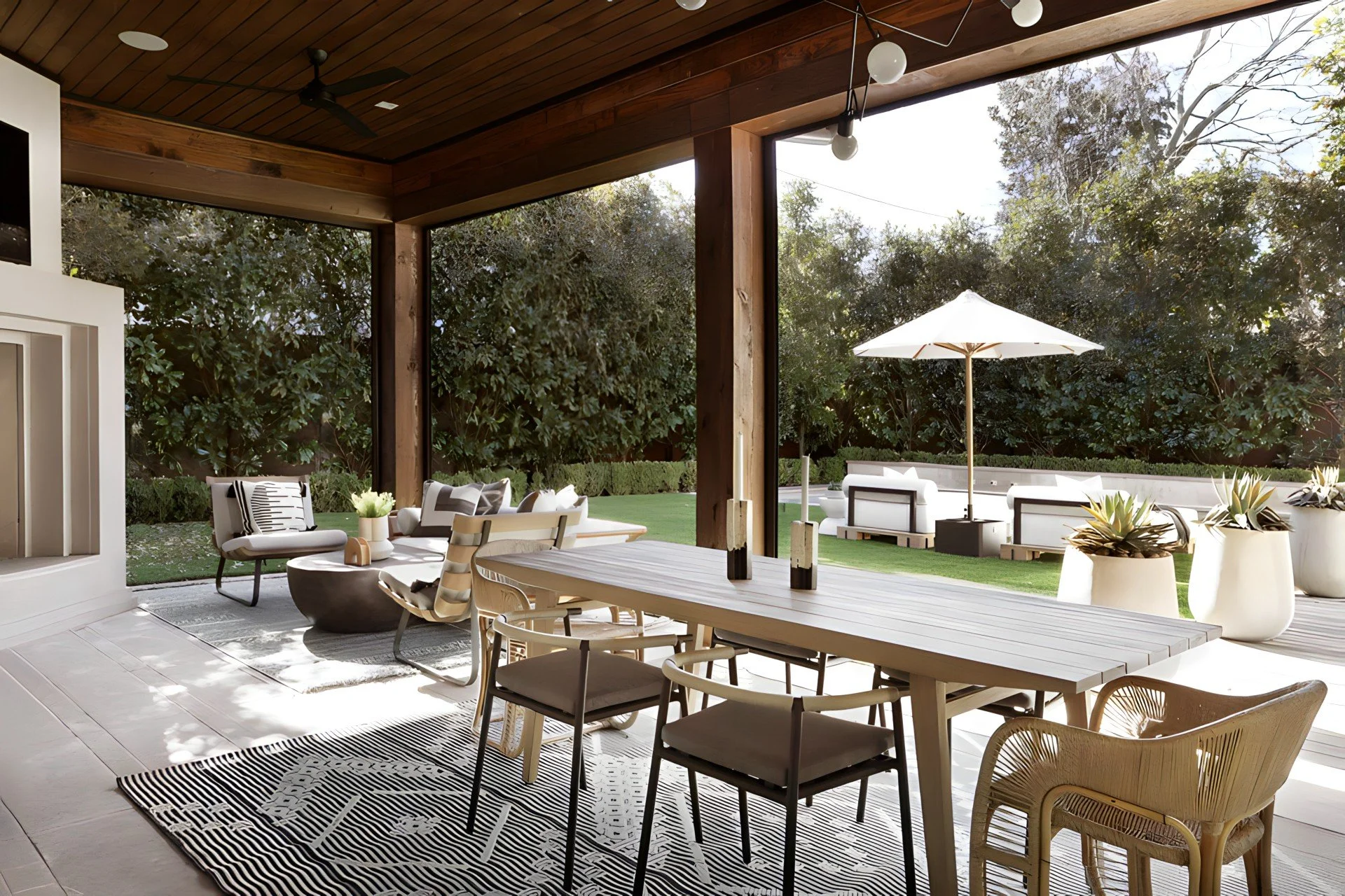 Indoor-outdoor living space with a wooden ceiling, a dining table with chairs, a sitting area with sofas and a coffee table, and a view of a lush garden with outdoor furniture and an umbrella.