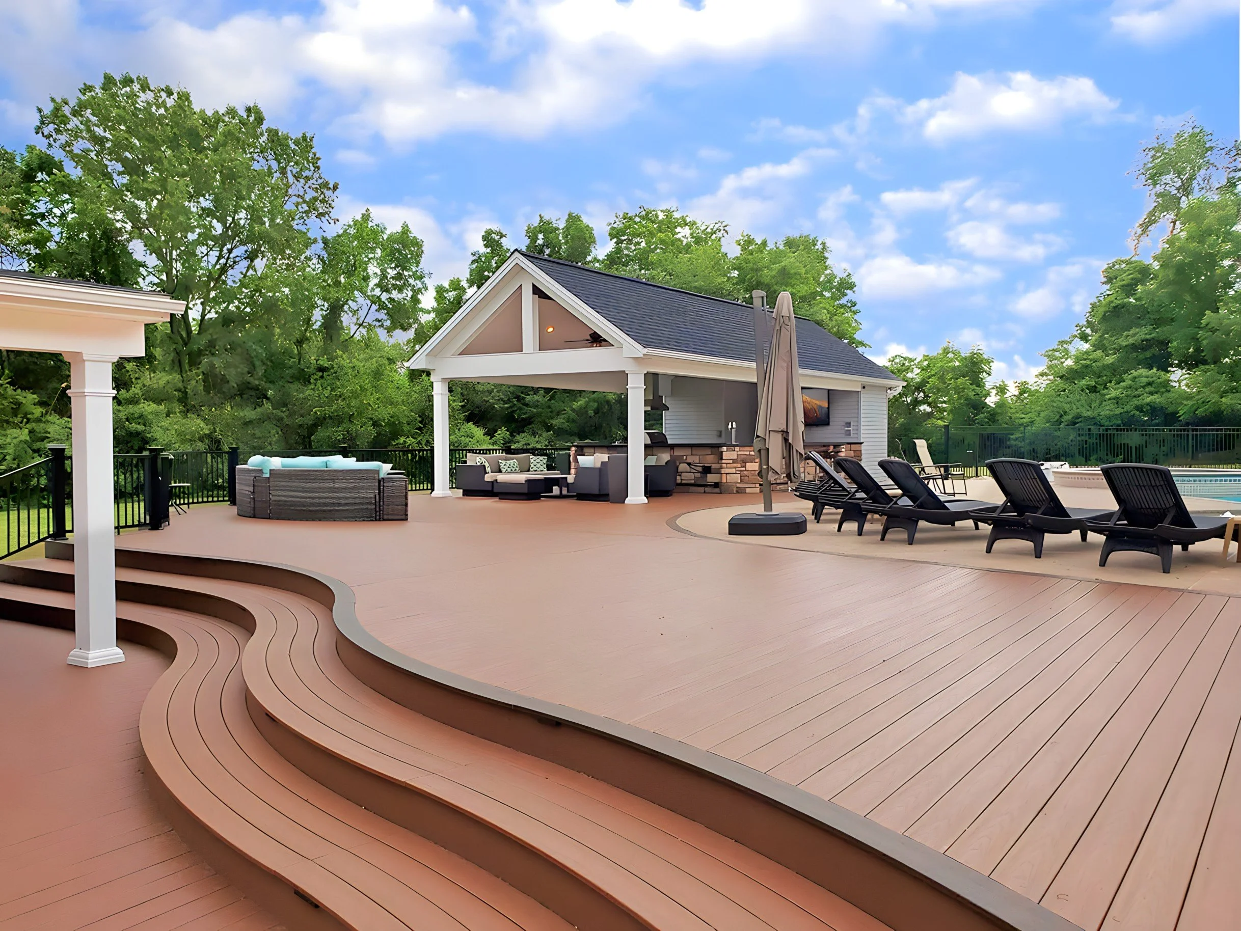 Spacious backyard patio with seating, lounge chairs, umbrella, and swimming pool, surrounded by green trees under blue sky with clouds.