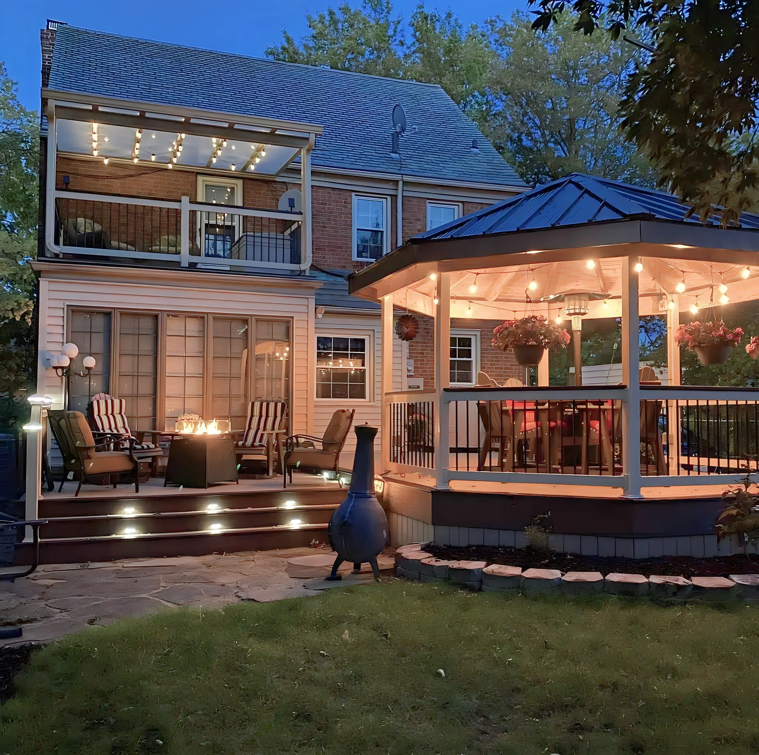 A two-story house with a brick exterior, decorated patio with a fire pit, outdoor seating, and a raised, lit gazebo with hanging flowers and string lights, all set in the evening. Trees are visible in the background.