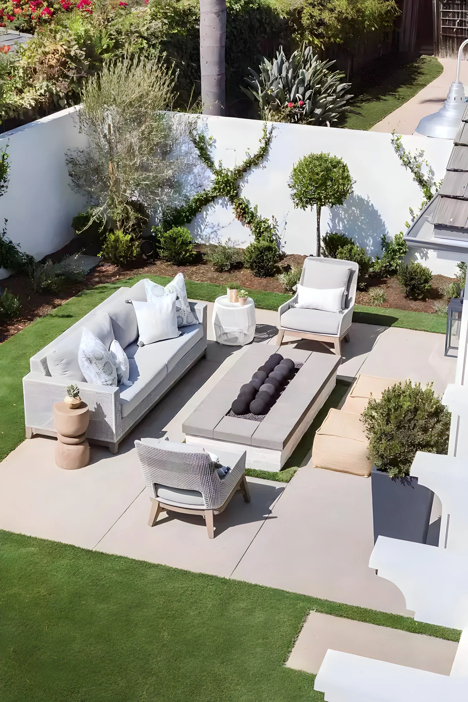 A modern backyard patio with white outdoor furniture, a central fire pit, potted plants, and a well-manicured lawn surrounded by a white wall and lush greenery.