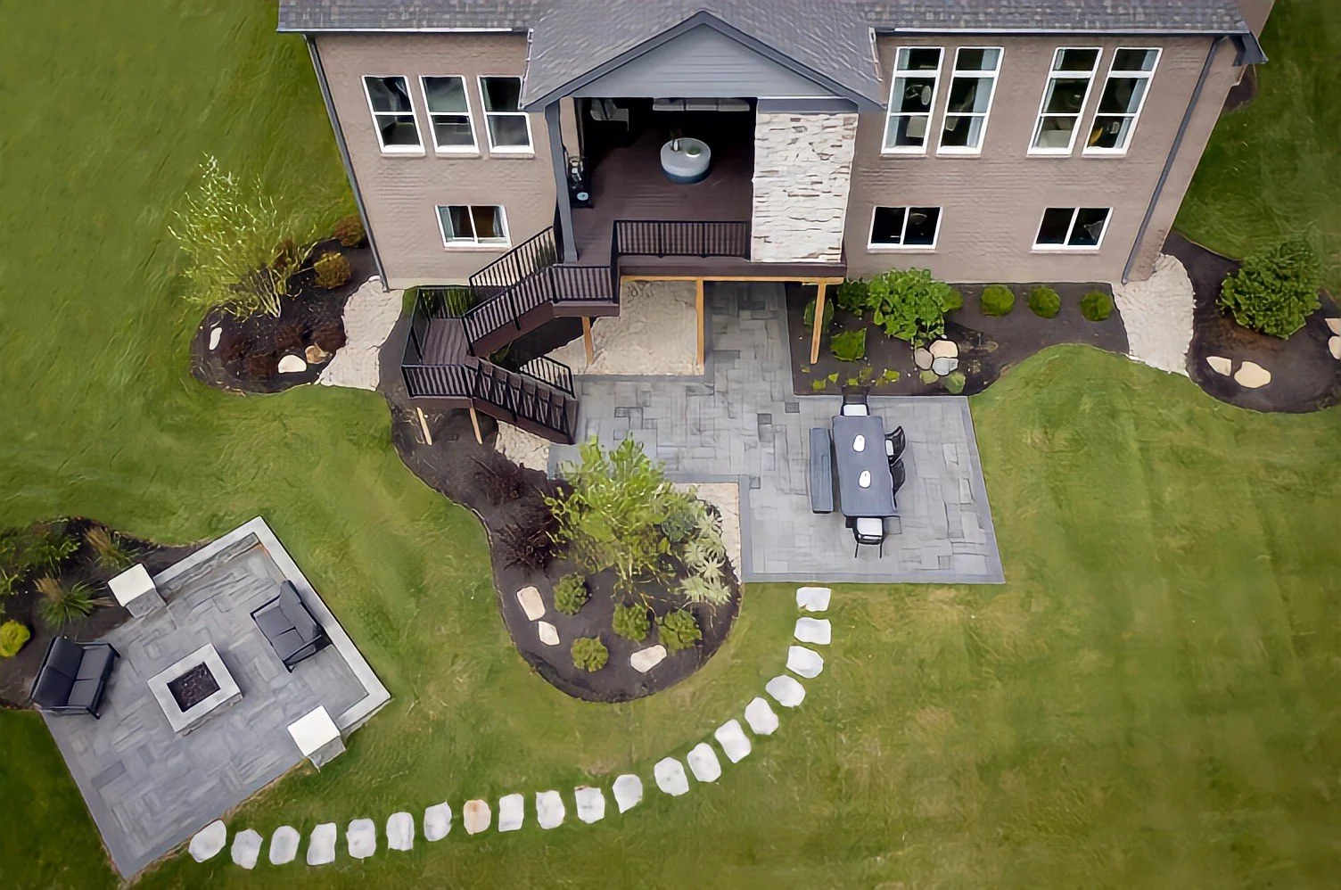 Aerial view of a backyard with a modern house, covered patio, outdoor dining table, fire pit area, landscaped garden beds, stepping stones, and a grassy lawn.