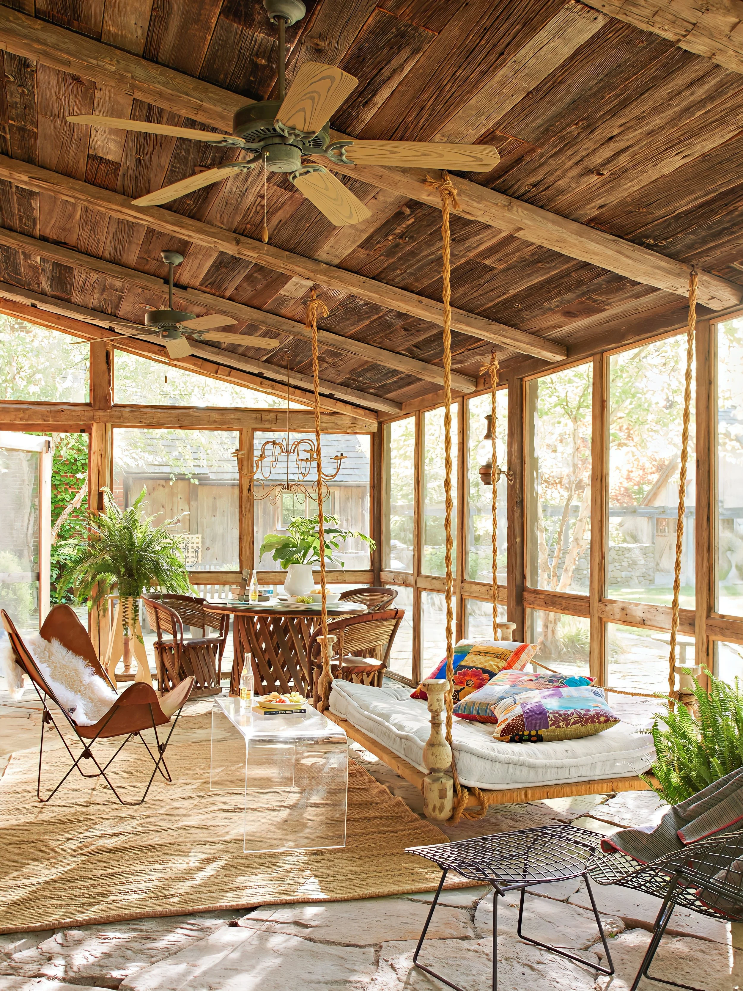 Bright sunroom with wooden ceiling and large windows, furnished with a hanging daybed with colorful pillows, a wooden dining table with chairs, a leather lounge chair with a fur pillow, and potted plants, illuminated by natural light.